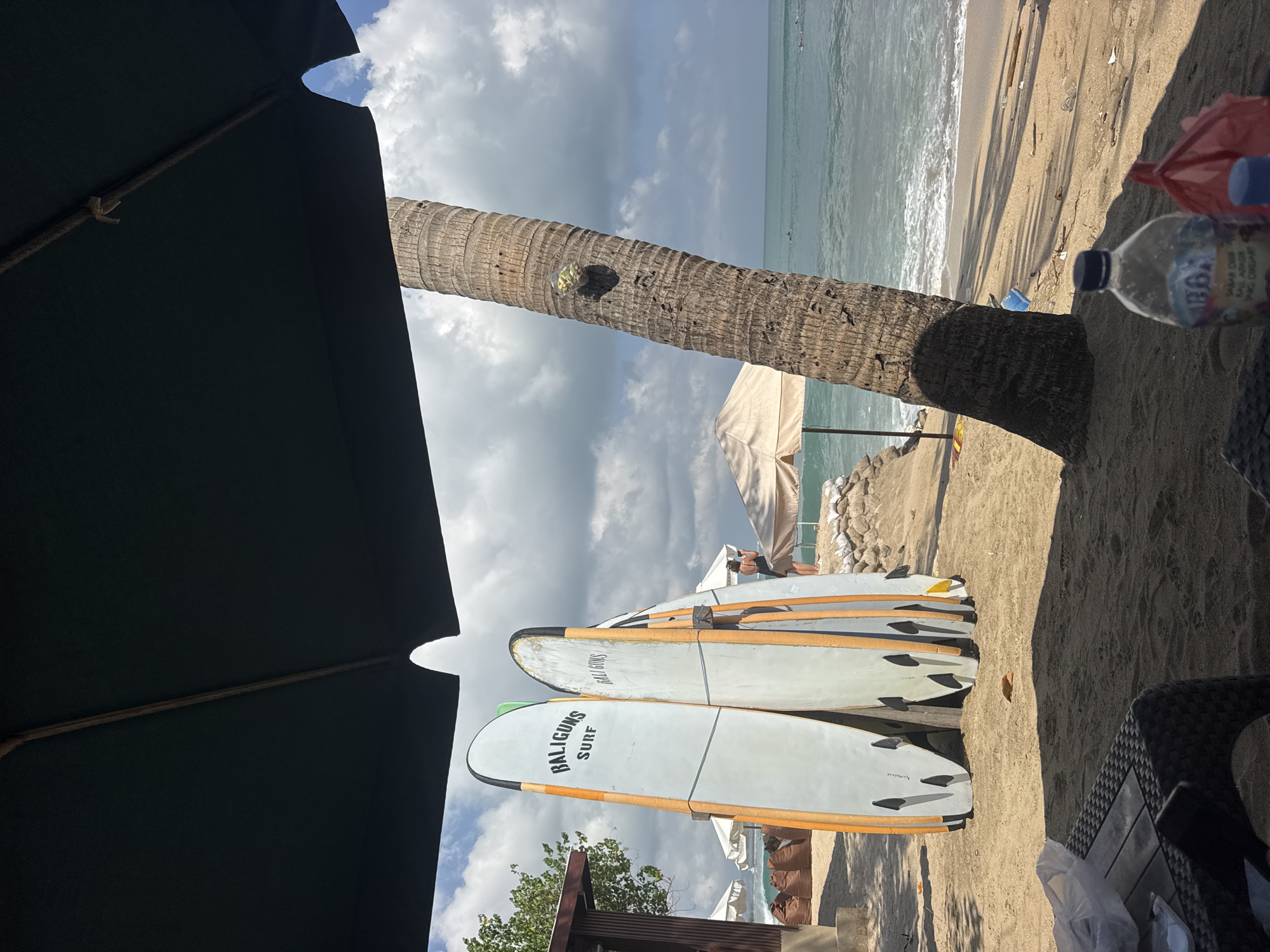 This is a photograph taken at Kuta Beach, Bali, Indonesia. The image is framed by a large dark umbrella, casting a shadow over the foreground. Two surfboards, branded "BALIGUNS SURF," are prominently displayed on the sand. To the right of the surfboards, a coconut palm tree stands tall, its trunk textured and weathered. A plastic bag filled with what appears to be fruit or snacks is attached to the tree. Beyond the palm tree, the turquoise waters of the ocean meet a sandy beach. Waves are gently rolling in, and a few people can be seen in the water, presumably surfing or swimming. Further down the beach, several beach umbrellas and beanbag chairs are visible, suggesting a relaxed atmosphere. The sky is partly cloudy, with a mix of white clouds and blue sky. In the foreground, a woven table and chair are partially visible, along with a plastic water bottle and a red plastic bag. The overall scene depicts a typical day at a tropical beach resort.