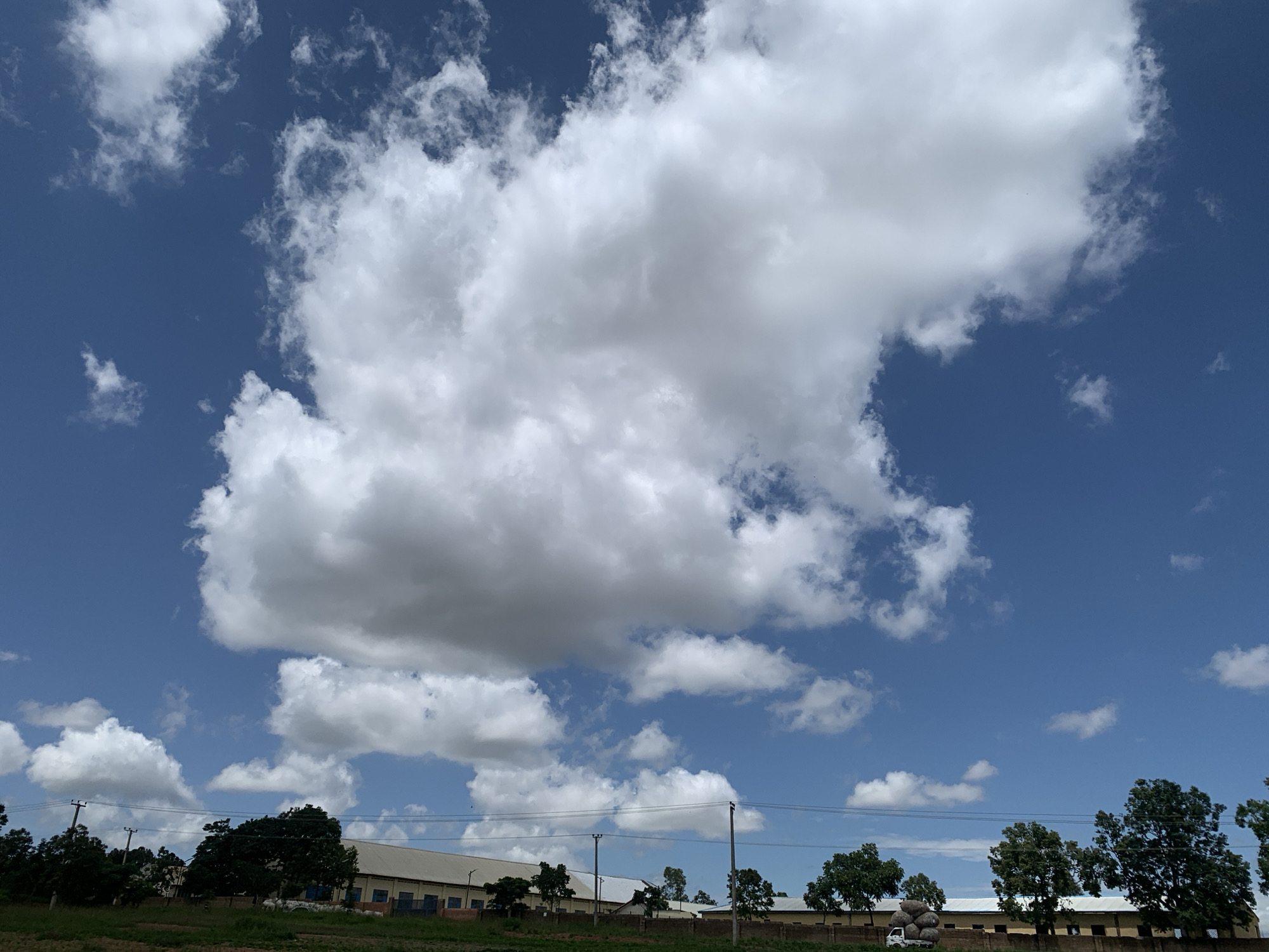 This is a wide shot of a rural landscape with a clear blue sky filled with fluffy white clouds. In the foreground, there is a patch of green grass and a low brown brick wall. Behind the wall, there is a long, yellow building with a white roof and a row of blue windows. Several trees, some with dark green foliage, are scattered throughout the scene. In the distance, there are utility poles with wires running between them. On the left side of the image, a white truck is parked near the building, loaded with large, dark, round objects. The scene appears to be located in Kuru Karama, Nigeria, possibly a factory or industrial area due to the long building and utility infrastructure. There are no people visible in the image. The overall impression is one of a bright, sunny day in a somewhat industrial rural setting.