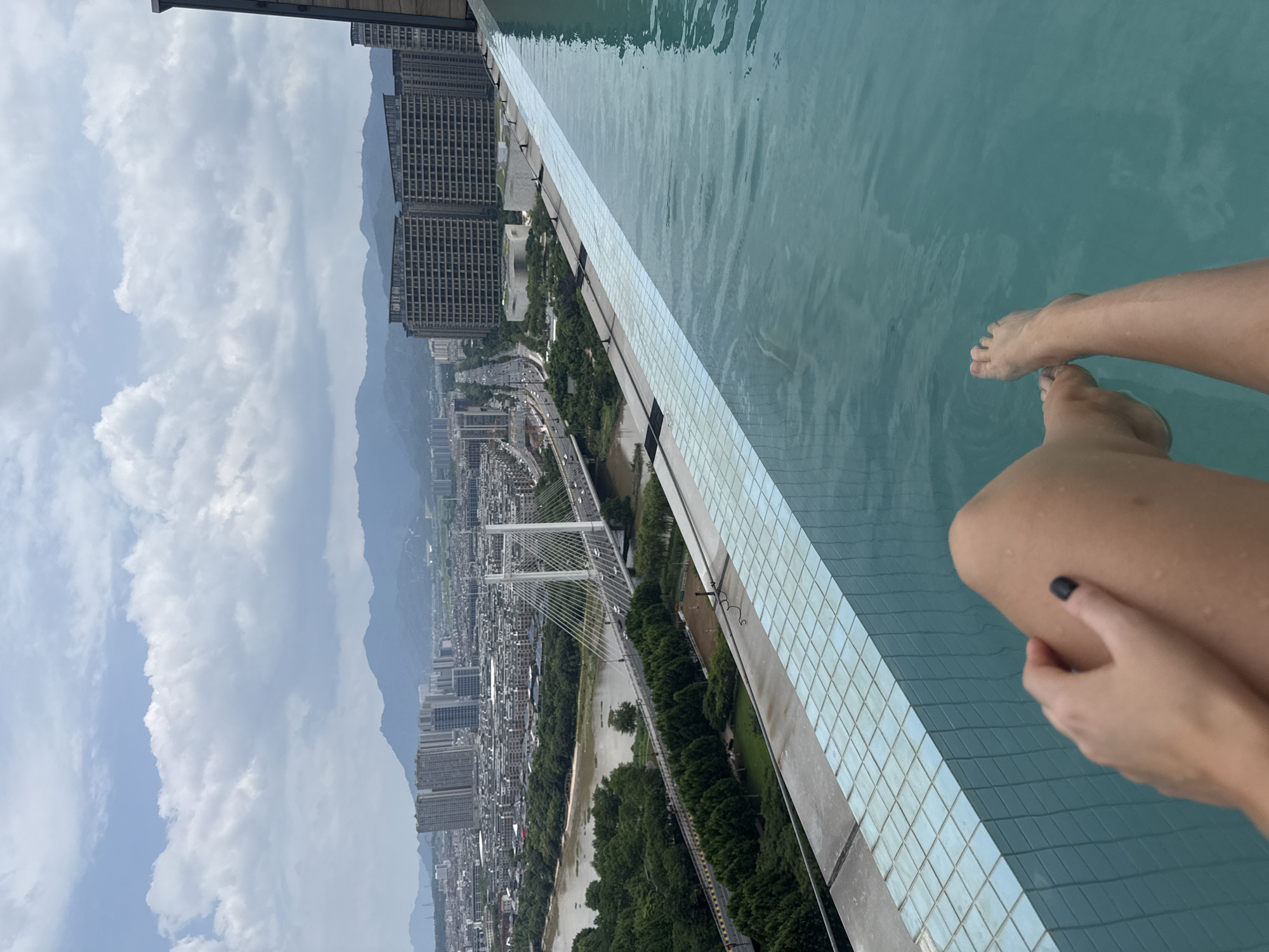 The image shows a person's legs and feet submerged in a turquoise infinity pool, with a sprawling cityscape of Yiwu, China visible in the background. The pool is bordered by a light blue tiled edge. Beyond the pool, a river winds through the city, spanned by a modern cable-stayed bridge with vehicles crossing. Numerous high-rise buildings and mountains form the urban and natural landscape under a cloudy sky. The perspective is from the pool looking out over the city.