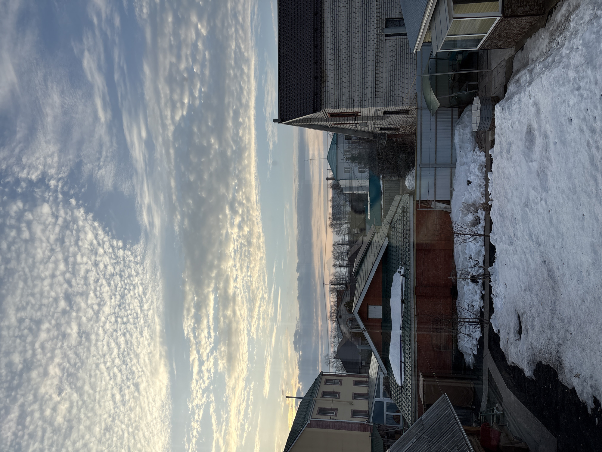 The image depicts a partially-overcast sky above a residential area in Ulyanovsk, Russia, possibly during late winter or early spring. There are layers of high-altitude cirrocumulus clouds with some lower-lying clouds. The horizon is visible with bare trees. Below are several houses with varying rooflines and brick or siding exteriors. Piles of melting snow are visible near the houses. There is no explicit presence of people, though it is implied that the residences are inhabited. The objects are houses, trees, and snow. Interactions are implied. The environment is a mix of winter-like conditions and a brightening sky.
