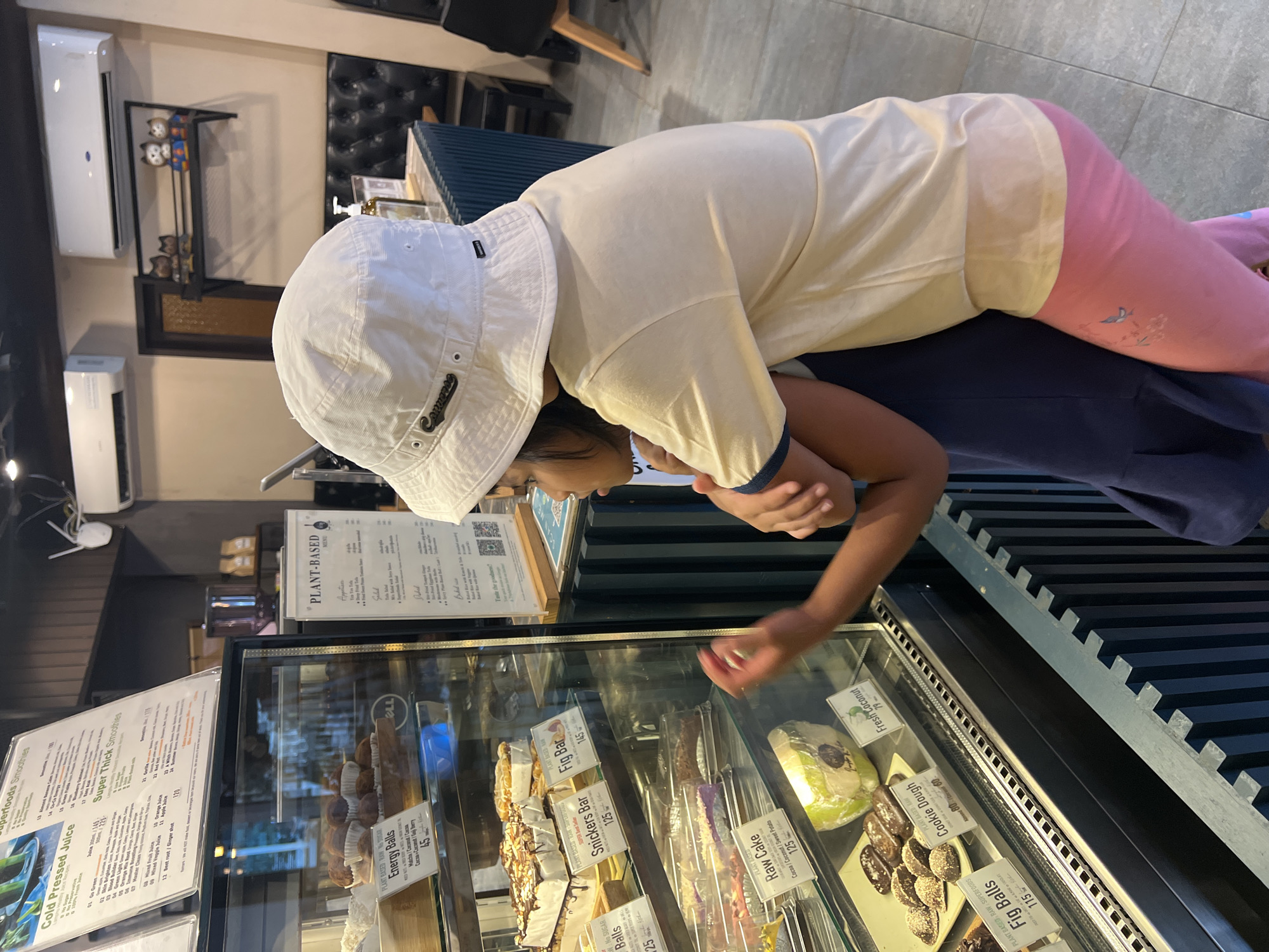 This indoor image captures a young child keenly observing a refrigerated display case filled with plant-based desserts and snacks. The child, dressed in a white bucket hat with "Converse" emblazoned on it, a light-colored t-shirt, and pink pants, is leaning over the glass case with their right hand extended, seemingly pointing at an item.
Inside the display, an assortment of treats is visible, each labeled with its name and price. Notable items include "Energy Balls" (45), "Snickers Bar" (125), "Fig Bar" (145), "Raw Cake" (125), "Fig Balls" (115), and "Cookie Dough" (80/60). A "Fresh Coconut" is also available for 79. Many labels specify "PLANT-BASED" and "RAW," indicating a focus on healthy or vegan options.
Menus are displayed adjacent to the case. One prominent menu is labeled "PLANT-BASED," listing appetizers and salads with the phrase "Taste the goodness!". Another menu advertises "Superfoods Smoothies" and "Cold Pressed Juice," featuring options like "Go Green," "Green Light," "Mixed Fruit Juice," and "Beetroot / Ginger Shot," along with their respective prices.
The background features white air conditioning units mounted on a light wall, a decorative black shelf adorned with owl figurines, and dark, possibly upholstered, seating. The floor is tiled. The child appears engaged and focused, likely selecting a treat in what appears to be a cafe or health food establishment in Baan Lamai, Thailand.