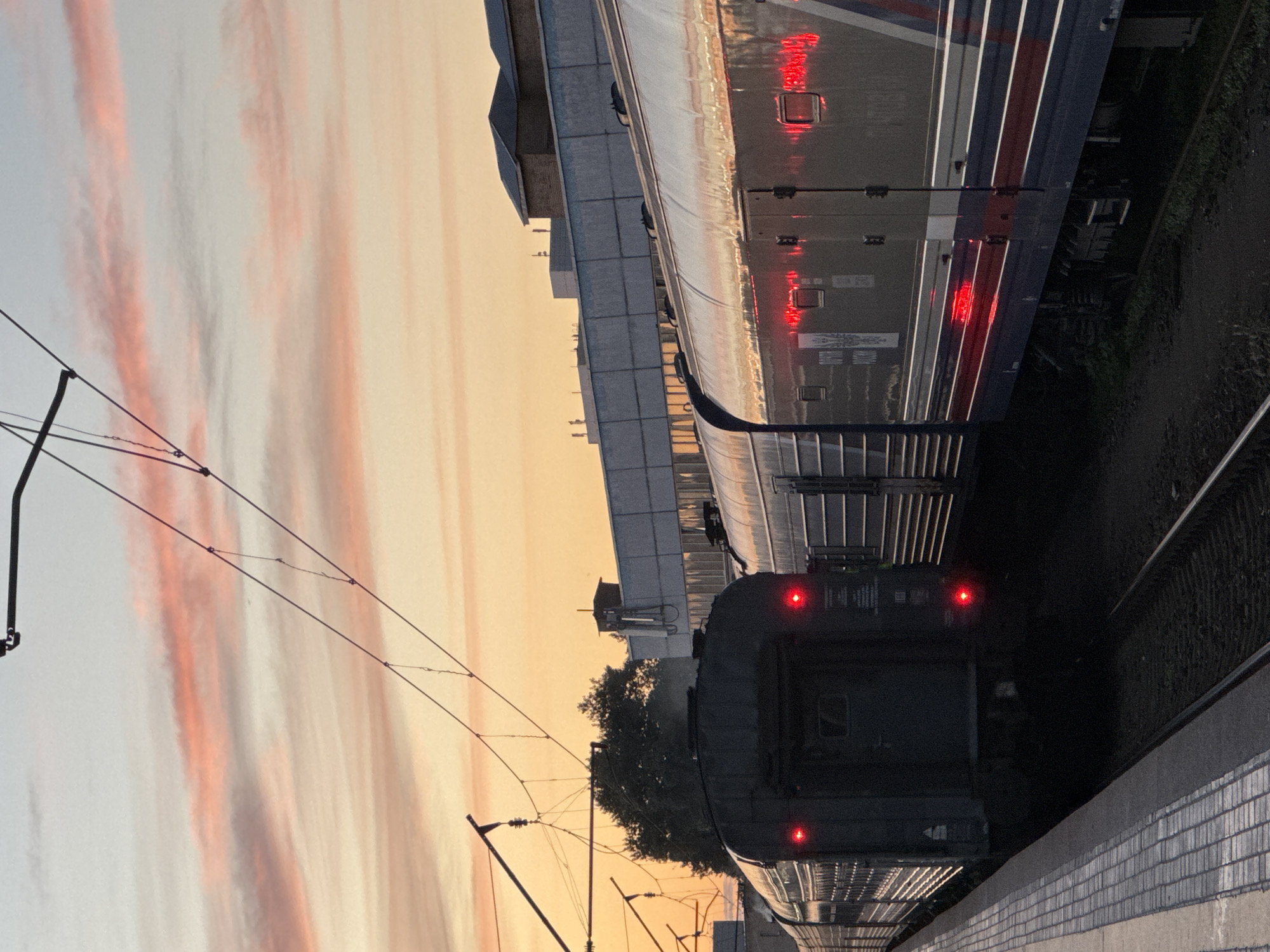 The image captures a train at a station during sunset, with the sky painted in hues of orange, pink, and yellow due to streaky clouds. Two trains are visible on adjacent tracks. The foreground train is dark and appears to be at the platform, with its red taillights illuminated. The second train, further down the track, is lighter in color and its windows reflect the sunset. The station platform has a tiled surface, and there are overhead power lines for the trains. The scene suggests a tranquil evening at a railway station, possibly in Moscow, Russia, as indicated by the context. No people are visible in the image. There is some text on the side of the lighter train, but it is not legible. The overall mood is peaceful, illuminated by the soft glow of the setting sun.