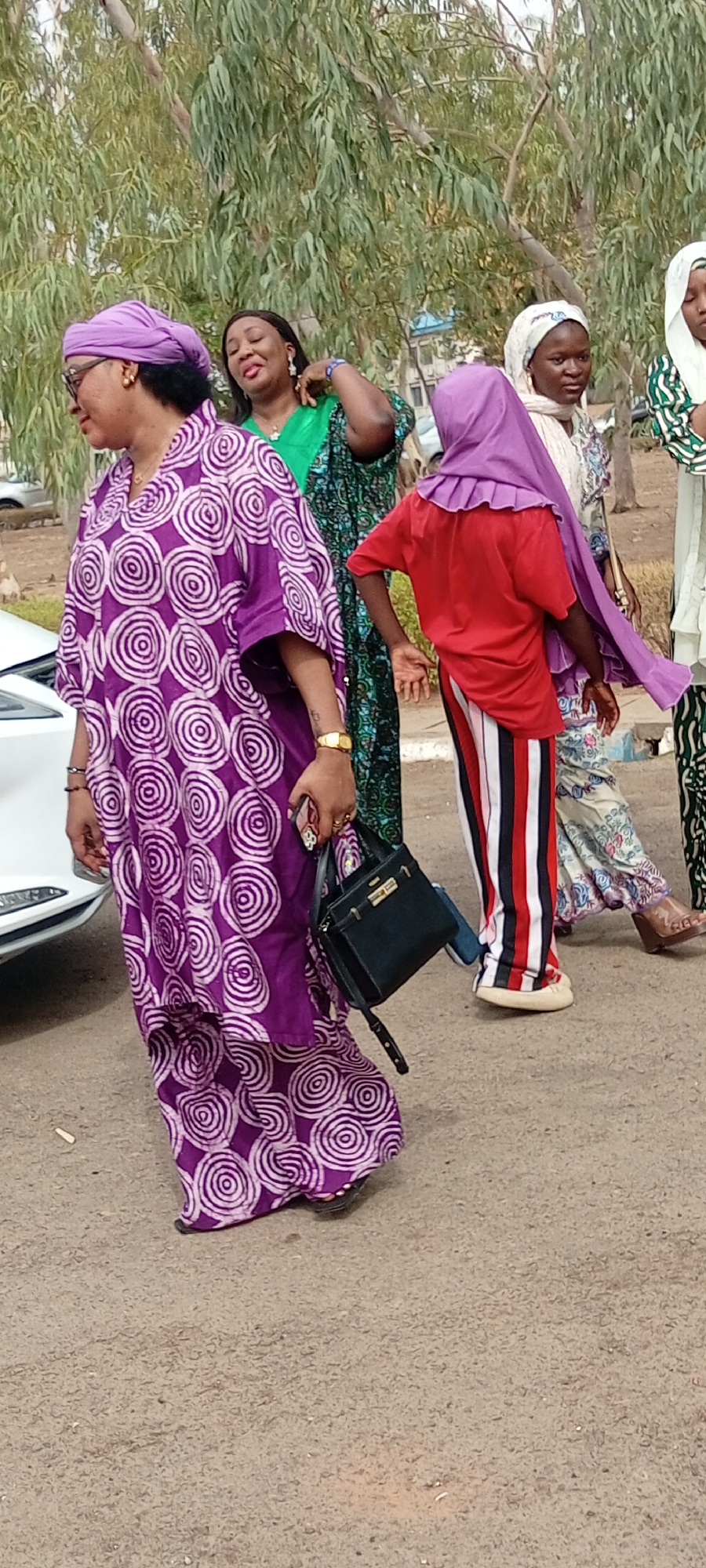 In Goron Dutse, Nigeria, a group of women walk on a paved surface. The woman in the foreground wears a purple patterned dress and matching headwrap, carries a black purse, and holds a phone in her hand. Behind her, a woman wears a green dress and jewelry. To the right, a person in a red shirt, purple head covering, and red and white striped pants stands partially obscured. Another woman in a patterned dress and white headscarf is seen in the background. Trees are visible in the backdrop. A portion of a white car is visible on the left.