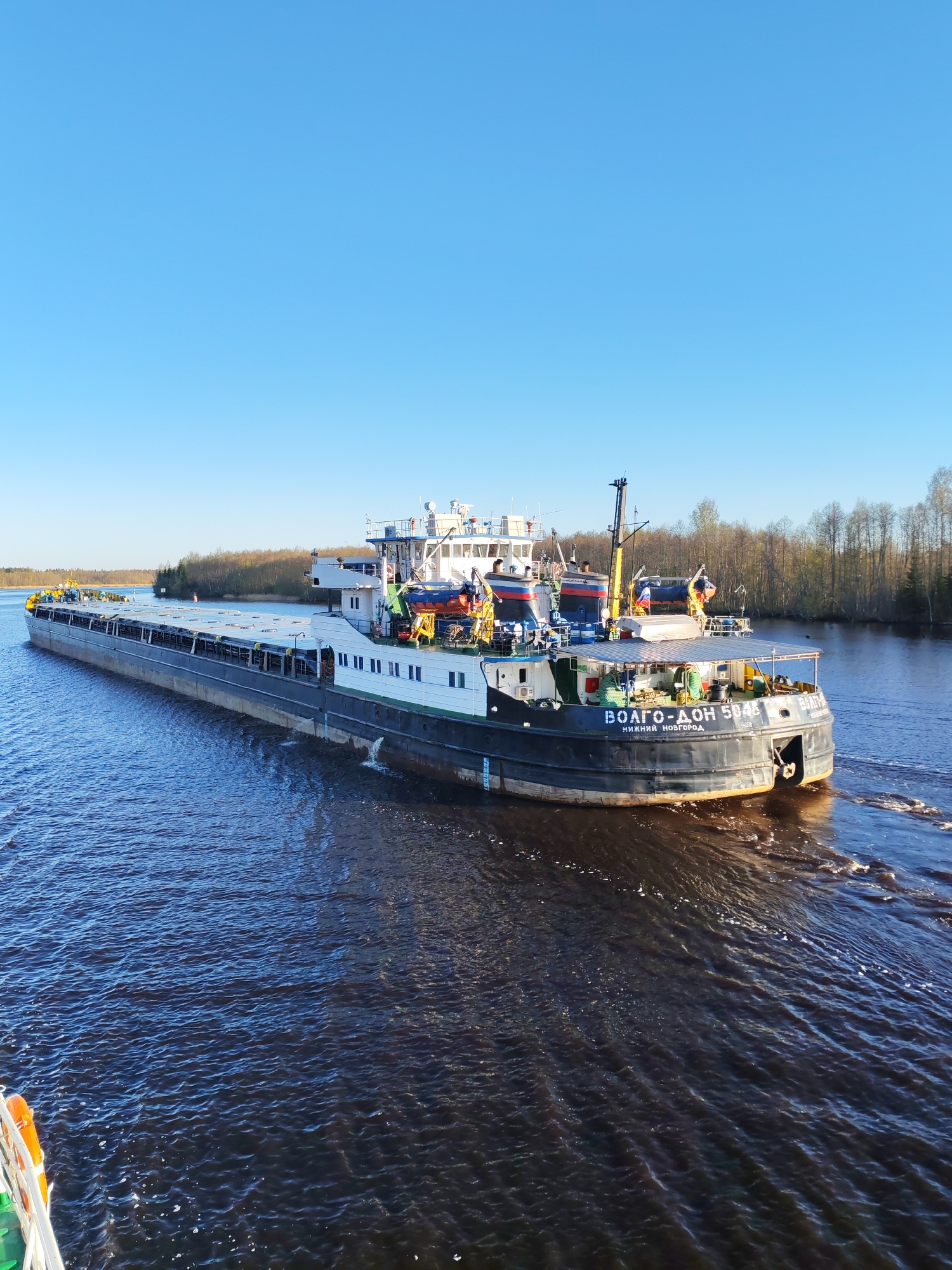 The image depicts a large river barge underway on a waterway in Novokemskii, Russia. The barge, identified as "VOLGO-DON 5046 NIZHNIY NOVGOROD" is dark-hulled with a white superstructure. It is moving from right to left in the frame, leaving a wake in the dark water. The sky is clear blue. The background consists of a forested shoreline along the riverbank. The perspective is from a slightly elevated viewpoint, possibly from another vessel or structure on the water.