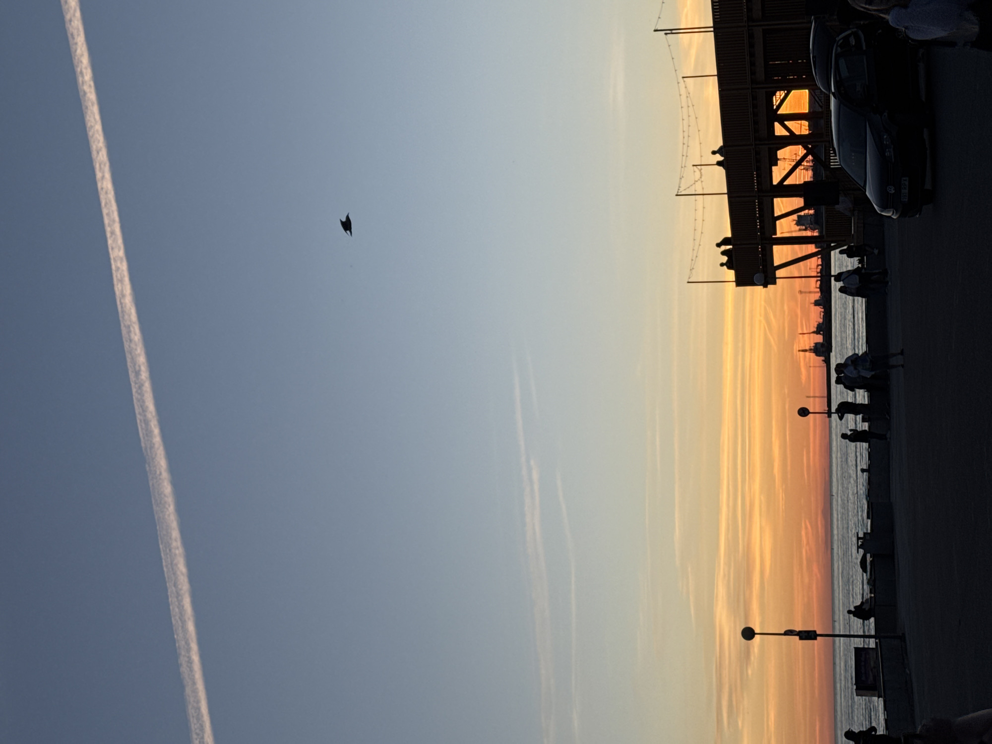 The image depicts a sunset scene in Tallinn, Estonia, likely near the harbor or waterfront. The sky transitions from a clear blue at the top to vibrant orange and yellow hues near the horizon, with wispy clouds and a prominent contrail stretching across the upper portion of the sky. A solitary bird is silhouetted against the sky.
In the mid-ground, a wooden structure resembling an elevated boardwalk or viewing platform stands silhouetted against the sunset, with several people visible standing on it. Below this platform, a body of water, likely the sea, is visible, with a faint outline of industrial cranes in the distance on the horizon.
In the foreground, a paved area is occupied by several silhouetted individuals, some standing, others seated. Two lampposts are present. A dark-colored car is parked on the right side of the foreground. The overall atmosphere is serene, capturing the end of the day with a focus on the natural beauty of the sunset and the silhouettes of human activity against it.