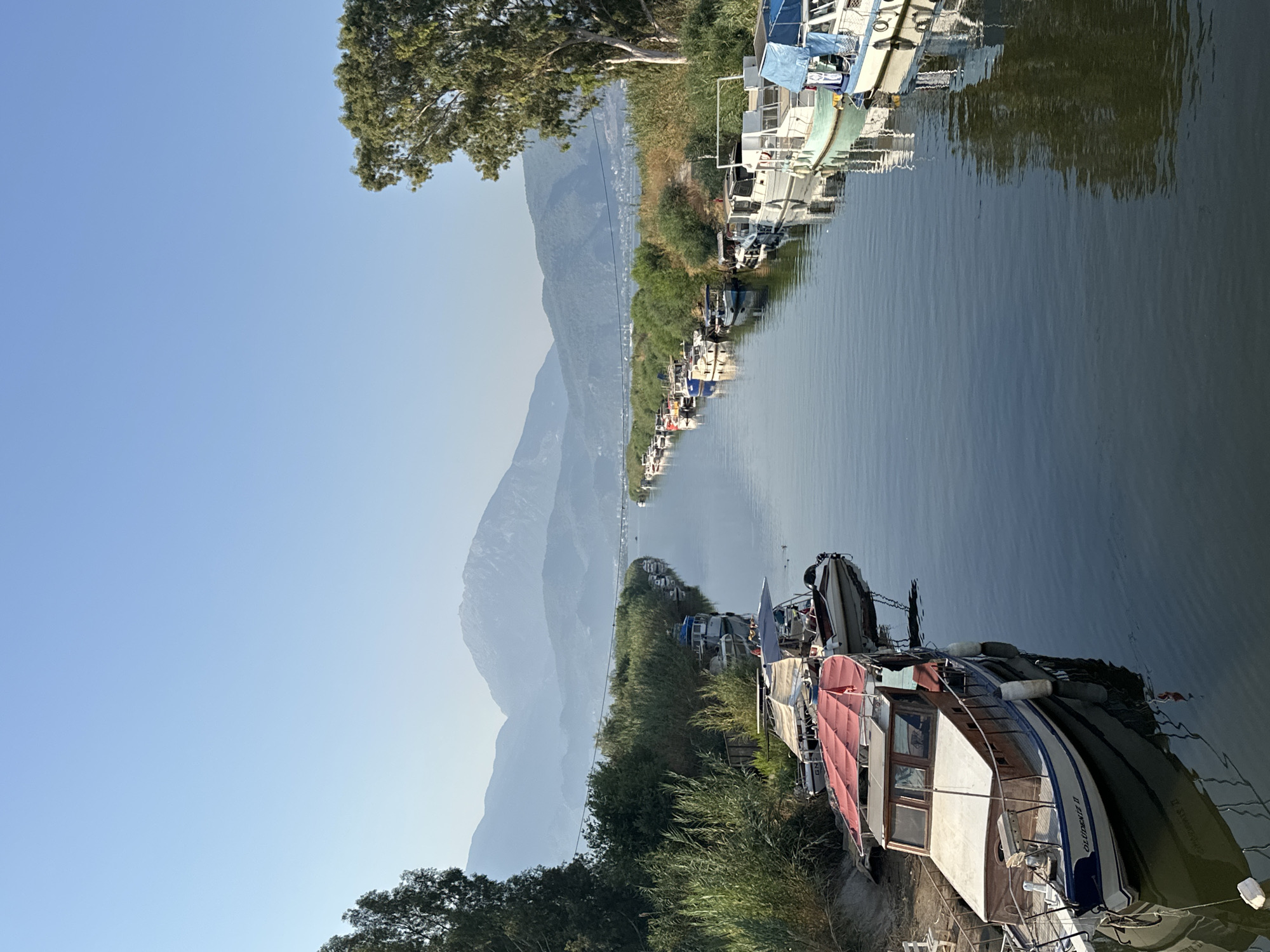 This is a photograph of a canal in Fethiye, Turkey. The canal is lined with trees and vegetation on both sides. Several boats are moored along the banks of the canal. The water in the canal is calm and reflects the surrounding scenery. In the background, mountains can be seen in the distance under a clear blue sky. The overall impression is one of a peaceful, natural setting within a town.