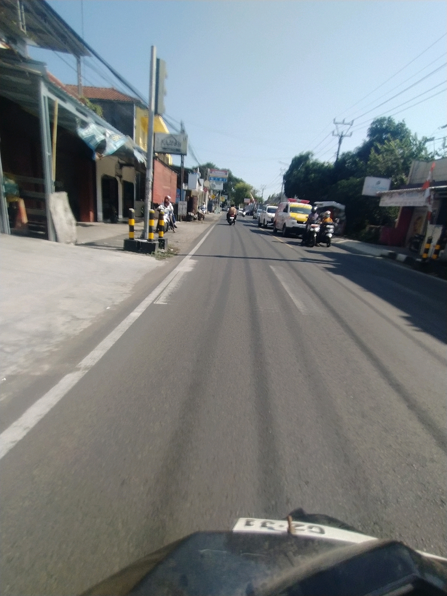 The image shows a view from a motorcycle traveling down a road in Desa Karangbenda, Indonesia. The road is paved and has a white line marking the lane. On the left side of the road are buildings with awnings and some parked motorcycles. On the right side, there are more buildings and vehicles including a yellow van that appears to be an ambulance, and several motorcycles. There are also utility poles and wires overhead. The sky is clear and blue. The scene appears to be during the daytime with bright sunlight casting shadows.