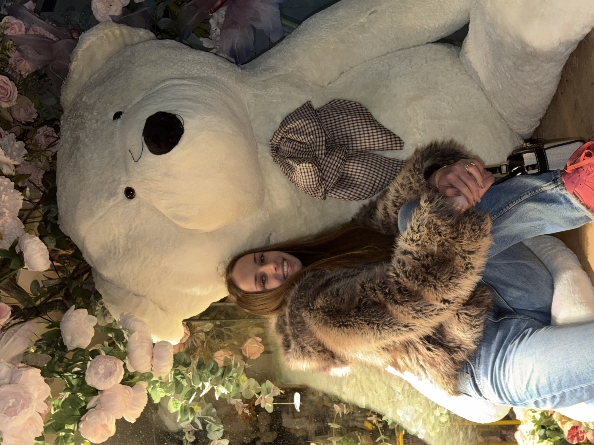 A young woman lies beside a large, white teddy bear in an interior space decorated with pink and white flowers. The bear has a checkered bow tied around its neck. The woman is wearing a fur coat and blue jeans, and her hands are clasped together. She appears to be smiling. A purse and pink sneakers are visible near her legs. The location is likely a shop or event space in Sofia, Bulgaria, given the context request. The scene seems to be set up for photographs.