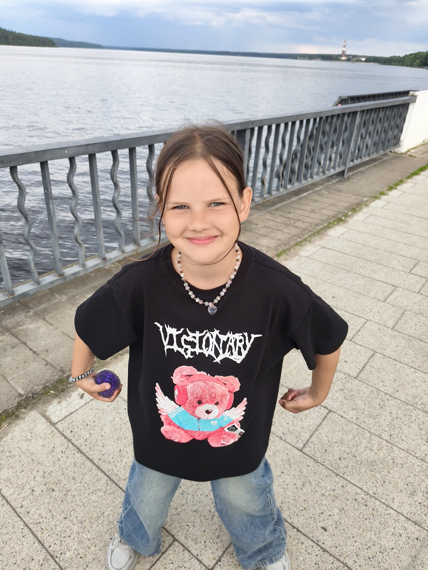 A young girl stands on a paved walkway next to a metal railing that borders a body of water. The girl is wearing a black t-shirt with a graphic of a pink teddy bear and the word "VISIONARY" in white lettering, blue jeans, and white sneakers. She is also wearing a necklace with clear and pink beads and a heart-shaped pendant, and a bracelet on her left wrist. In her left hand, she holds a small purple ball. The background shows a wide body of water, a distant treeline, and a cloudy sky. In the distance, a lighthouse-like structure is visible. The scene is likely set in the city of Chaikovsky, Russia, given the presence of the Kama River, which flows through the city, and the characteristic architecture of the area.