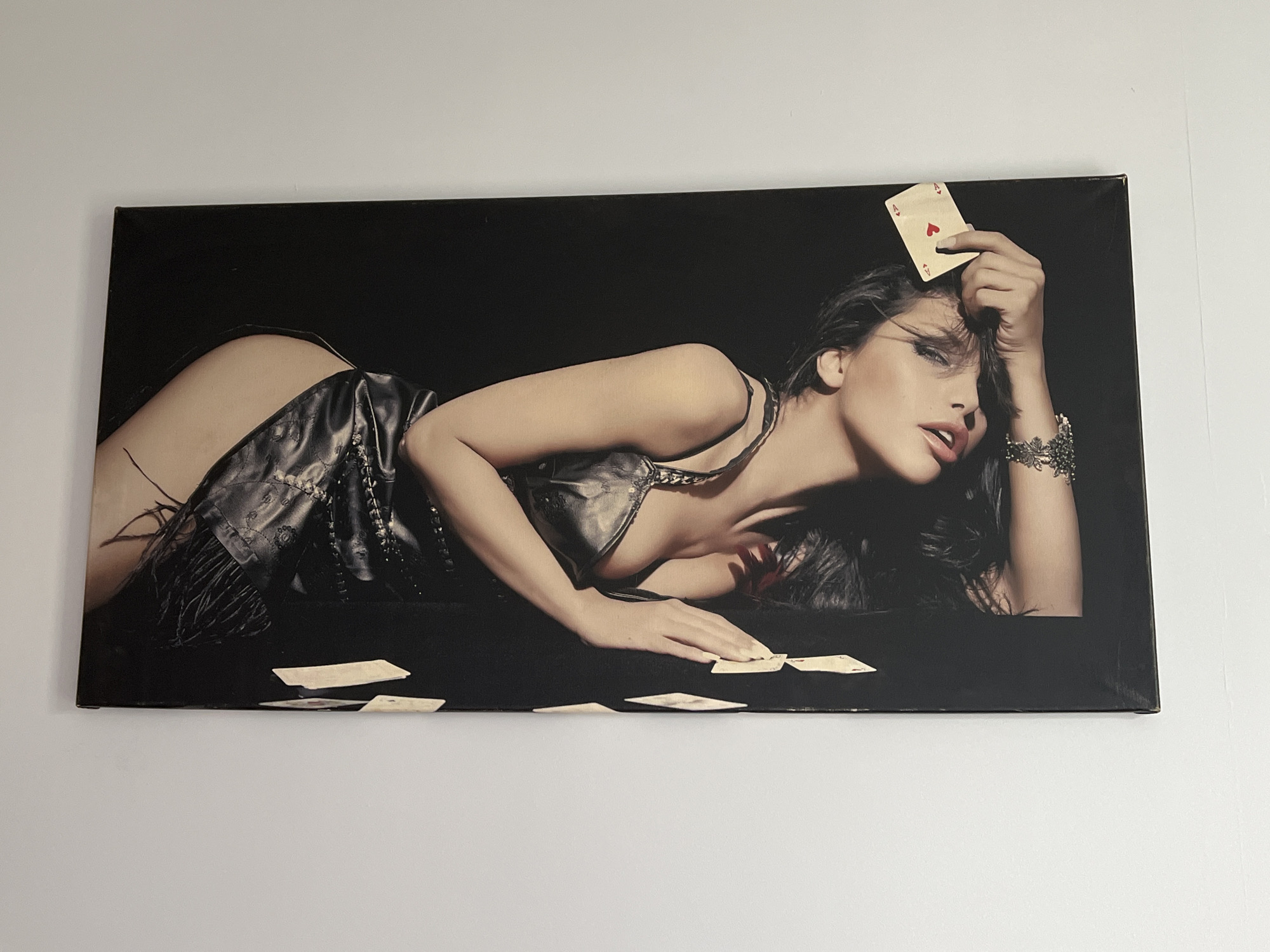 A horizontal canvas print depicts a woman lying on her side against a black background. She is wearing a black, strappy top and a black skirt or shorts with fringe detailing. Her left arm is bent, with her hand resting near some scattered playing cards on the surface. In her right hand, she holds an ace of hearts card up to her forehead. Her head is tilted back, and her eyes are looking forward, with her lips slightly parted. Scattered playing cards are visible on the black surface around her. The canvas print is mounted on a white wall.