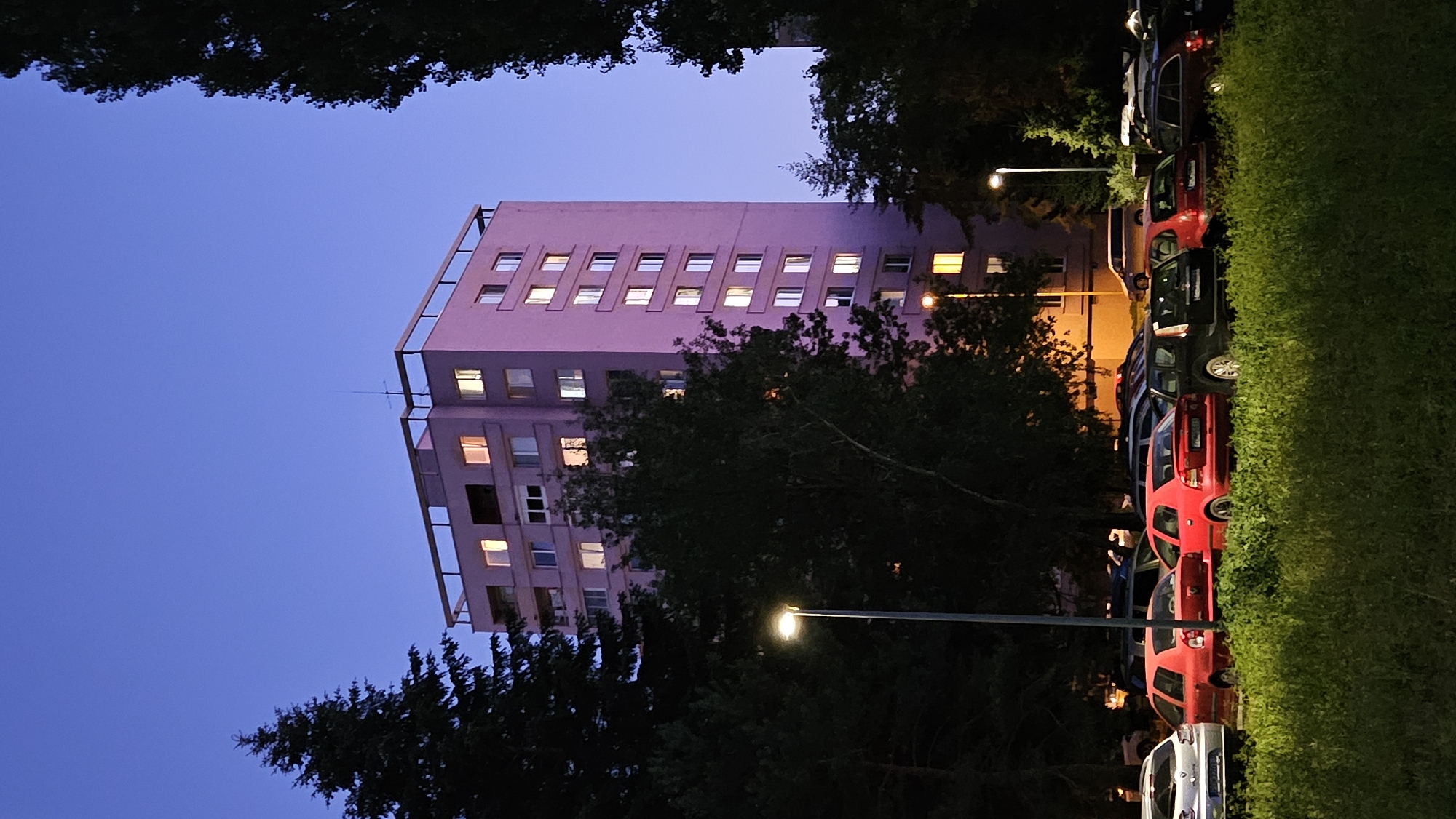 The image captures a twilight scene in the city of Prešov, Slovakia, with a tall, pink apartment building as the central subject. The building features numerous windows, many of which are illuminated from within, suggesting occupancy. The sky above is a deep twilight blue, with a few faint stars visible.
In the foreground, a parking lot is filled with various cars, including red sedans, a silver hatchback, and a black SUV. The cars are parked haphazardly, indicating typical urban parking conditions. A patch of lush green grass and foliage lines the bottom of the frame, partially obscuring the lower portions of the parked vehicles.
Tall trees, some dark and silhouetted against the sky, frame the apartment building and the parking lot, adding a sense of depth and natural elements to the urban landscape. Streetlights are visible, their illumination contributing to the overall ambiance of the scene. The image appears to be taken from ground level, looking upwards towards the apartment building. There are no people visible in the image. The overall impression is one of a quiet residential area at the close of day.