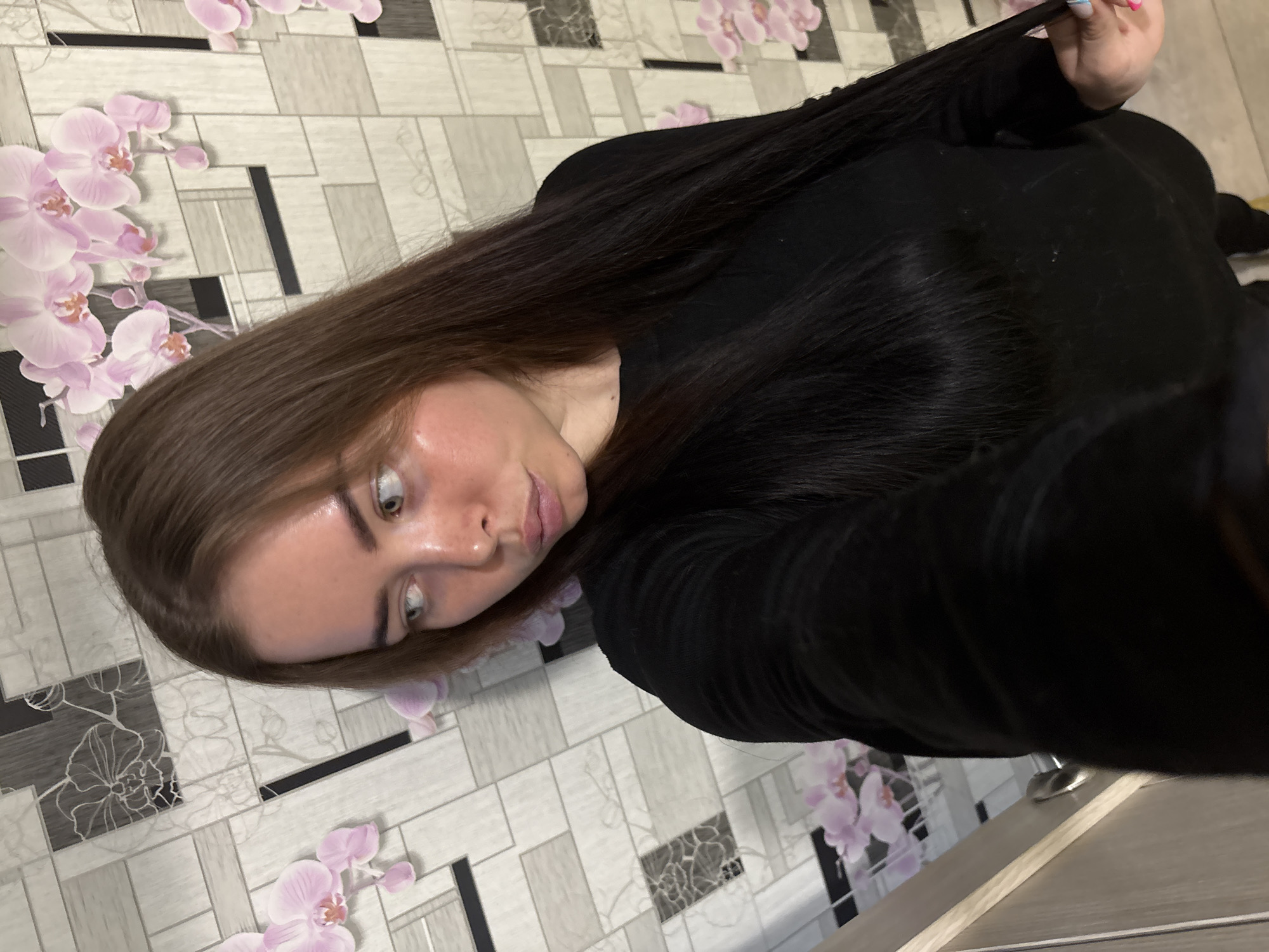 A young woman with light skin, brown hair, and light-colored eyes is posing for a selfie. She is wearing a black top. Her hair is long, dark, and flowing. In her right hand, she is holding a strand of her hair. The background is a wall with a pattern of tiles decorated with stylized orchid flowers. The lower right corner of the image shows the edge of what appears to be a wooden surface, possibly a table or furniture. There is no information in the image to determine that the picture was taken in Izhevsk, Russia.
