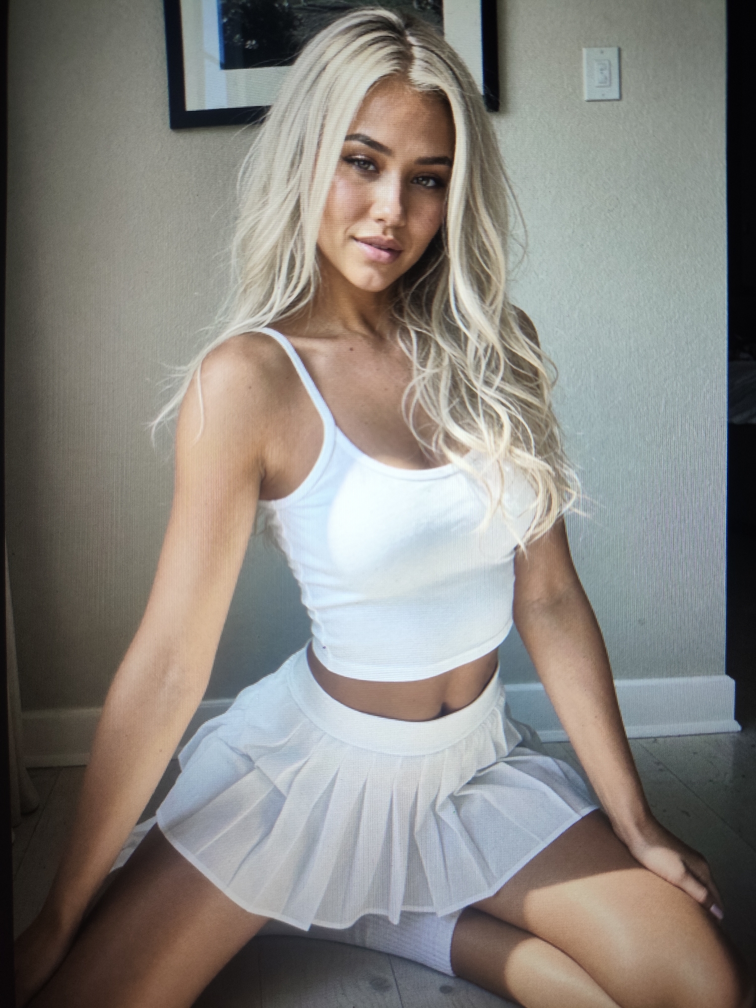 A fair-skinned female with long, wavy blonde hair, light eyes, and defined eyebrows is depicted looking directly at the viewer. She is wearing a white ribbed crop tank top with thin straps, a white pleated mini-skirt, and partially visible white knee-high socks. The individual is seated on a light-colored floor, with her hands resting on her thighs. The background features a plain, light beige wall. A dark-framed picture is partially visible in the upper left, and a white light switch plate with two switches is mounted on the upper right. The lighting is soft, casting subtle shadows on the floor. The scene is an unspecified indoor setting.