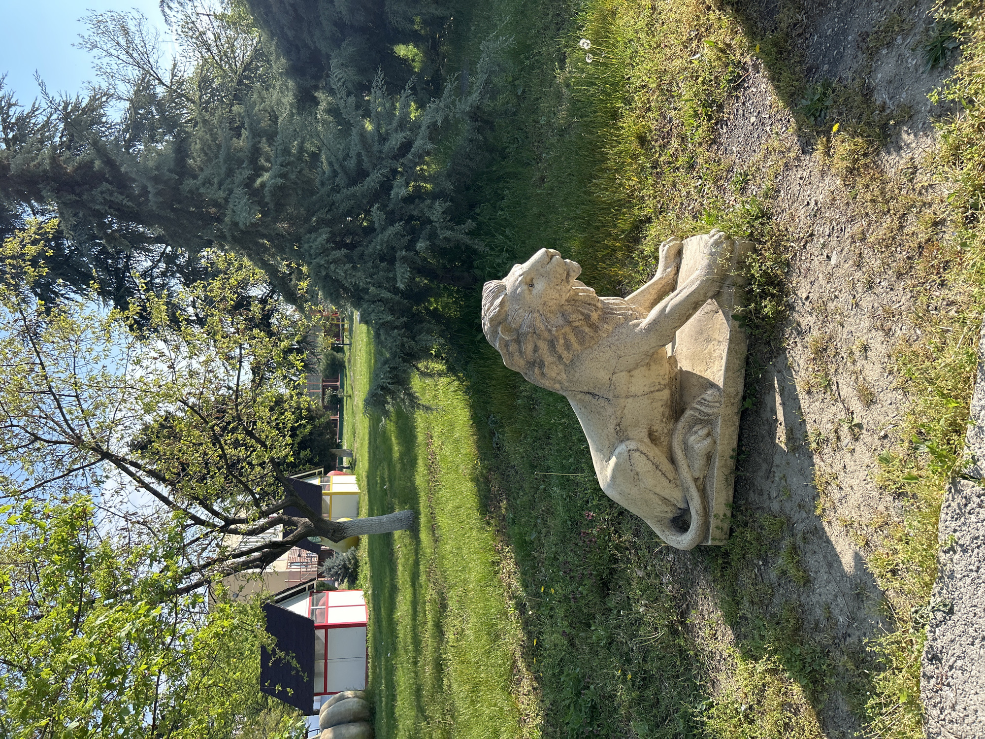 A stone lion statue stands on grass next to a path in a park-like setting. Trees of varying types surround the statue. In the background are buildings with red and white sections. The location is Plovdiv, Bulgaria.