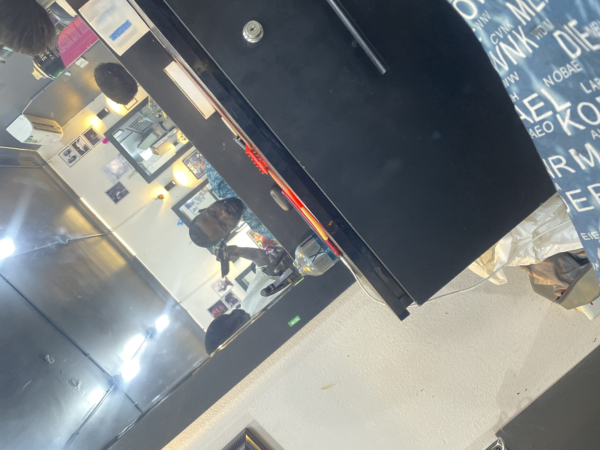 The image shows the interior of a barbershop, likely in Abuja, Nigeria, given the provided context. A dark-haired man is visible in the mirror's reflection, presumably a barber, holding a hair clipper, indicating he is likely cutting hair. The reflection reveals a wall decorated with framed pictures. The shop is equipped with a mirrored station and barbering tools. The surrounding environment appears to be a simple, functional space with white walls and a black cabinet or countertop. The lighting suggests it is either daytime or a well-lit space. A travel bag and other items are visible near the cabinet. The scene captures the essence of a typical barbershop setting.