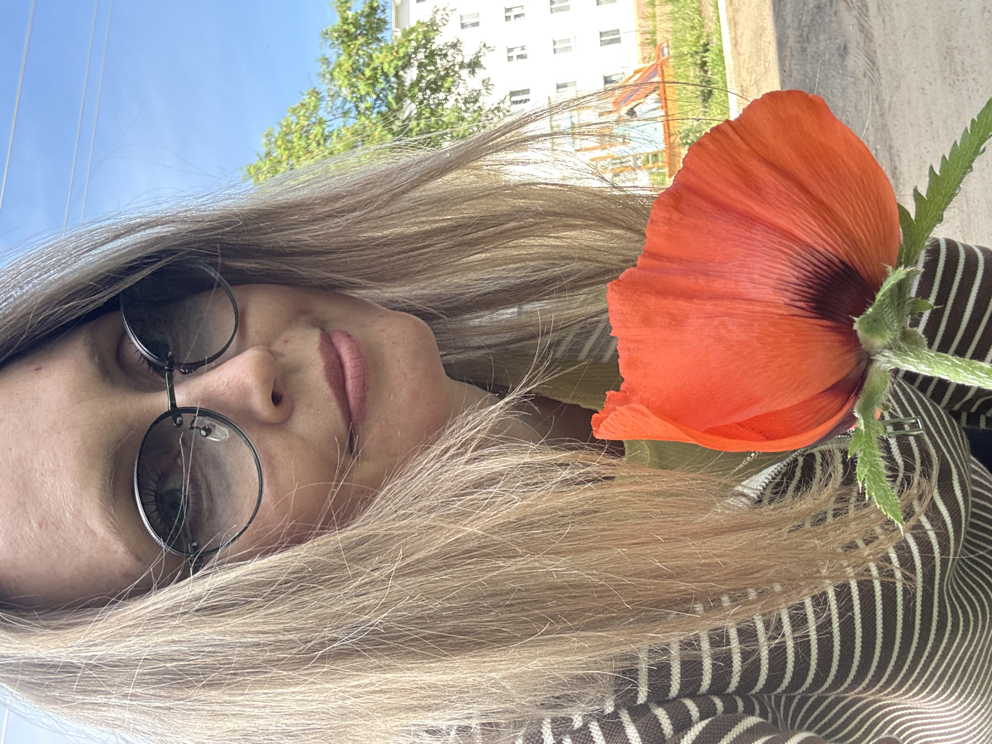 A blonde woman wearing sunglasses and a brown and white striped zip-up hoodie is holding a large red poppy in front of her face. She is outdoors on a sunny day. In the background, there is a white building with windows, a playground with slides, green trees, and a clear blue sky with power lines. The image appears to be taken in a residential area, possibly in Chaikovsky, Russia.