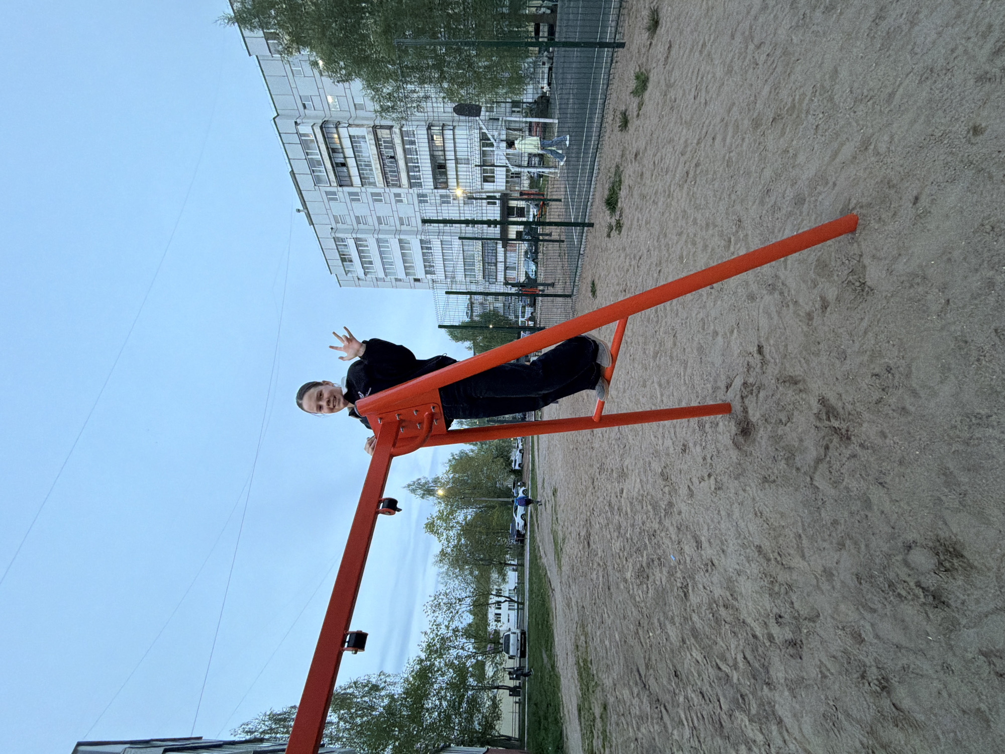 A rotated, long shot of a young person sitting on an orange playground ride in Chaikovsky, Russia. The person, dressed in dark clothing, looks at the camera and gives a peace sign. The background includes a light-colored apartment building, green trees, and a sand-covered playground area. Other playground equipment is visible behind a green fence. The sky is overcast and dusk is approaching.