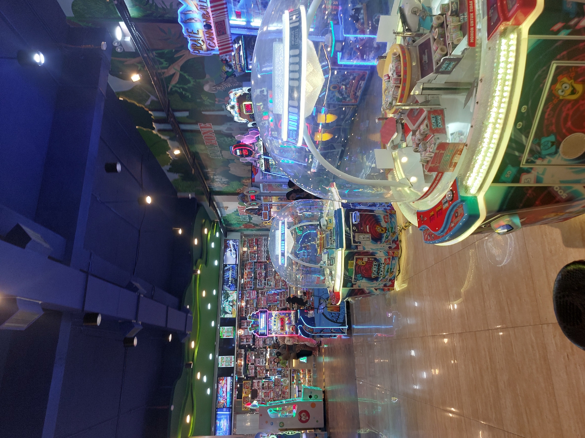 This image depicts the interior of an arcade in the City of Pematang Siantar, Indonesia. The arcade is brightly lit with a variety of colorful arcade games, many of which feature neon lights. In the foreground, a large, dome-shaped prize redemption game with a yellow top and red accents is prominent. The game has a clear plastic dome enclosing a display of tickets and other prizes. Behind this game, other arcade machines are visible, including a "Ticket Dome" machine and a brightly lit upright game with a cartoon character on it.
Further in the background, a wall is decorated with a mural depicting a jungle scene, complete with trees, animals, and text that reads "AMAZONE" and "PIRATE CANNONBALL". Several large screens are mounted on the wall above the shelves of prizes, displaying various graphics and games. A few people are seen in the background, appearing to play games or observe the machines. The floor is tiled with a polished, light-colored material that reflects the ambient light and the colorful glow of the arcade machines. The ceiling is dark blue and appears to have exposed ductwork and light fixtures. The overall atmosphere is one of a lively and engaging entertainment space.