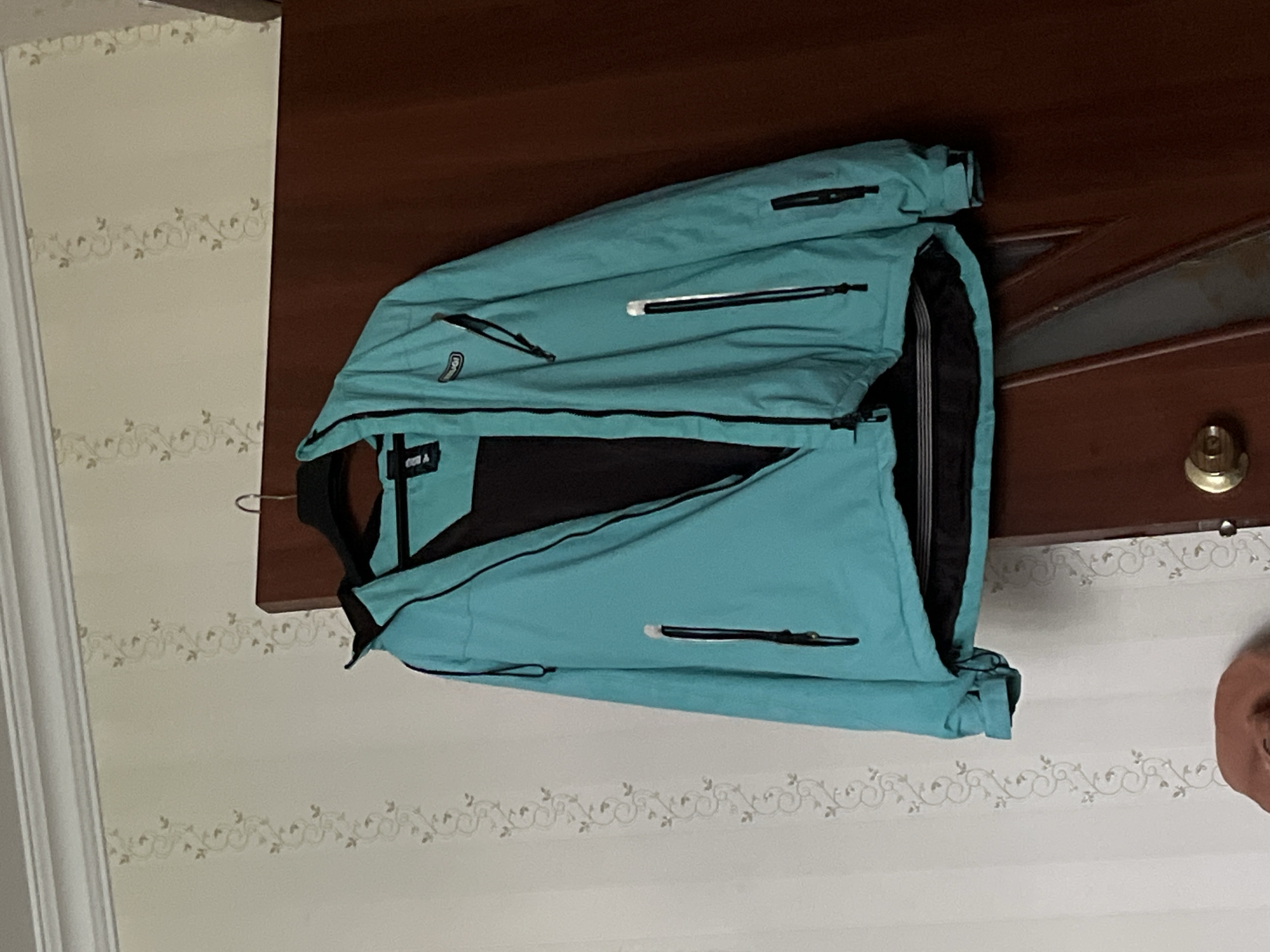 The image presents a teal-colored waterproof jacket, unzipped to expose its black inner lining, suspended on a black clothes hanger. The jacket features a central black zipper, two upper chest pockets with black zippers, one zippered pocket on the left sleeve, and two lower zippered pockets on the front. A partially visible brand tag, appearing to read "AUGI," is present near the collar area. The jacket's bottom hem reveals an internal black and white striped elastic band. The hanger is hooked over the top edge of a dark reddish-brown wooden door, which forms the background on the right side of the frame. The door exhibits paneling and a brass-colored round doorknob is visible in the lower right, alongside a reflective or glass panel insert. The left side of the image depicts a wall covered in light cream-colored wallpaper featuring vertical stripes of a scrolling vine and leaf pattern in a muted beige. A white decorative crown molding runs along the top edge of the wall. A small, indistinct reddish-brown fabric object is partially visible in the bottom left corner. No individuals are present.