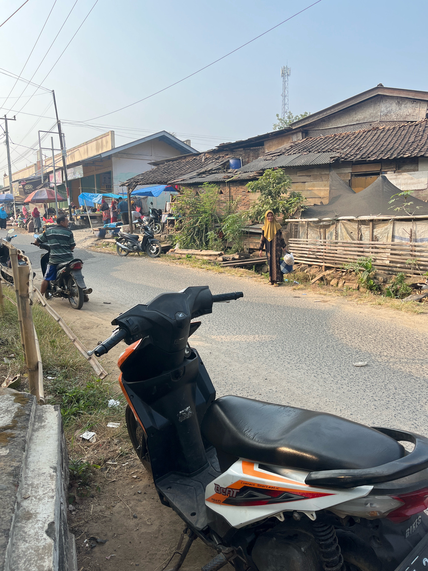 The image depicts a street scene in Margodadi, Indonesia, during daylight. A variety of motorcycles are parked and in use along a paved road lined with buildings and stalls, suggesting a marketplace or a busy thoroughfare. In the foreground, a black and orange scooter is prominently positioned. Further down the road, several people are visible, some tending to stalls under blue and red awnings, while others are on motorcycles. A woman wearing a yellow hijab and a long dress walks along the side of the road carrying bags. The buildings appear to be simple structures with tiled roofs, and some have corrugated metal. Vegetation is present along the roadside. A telecommunications tower can be seen in the distance.