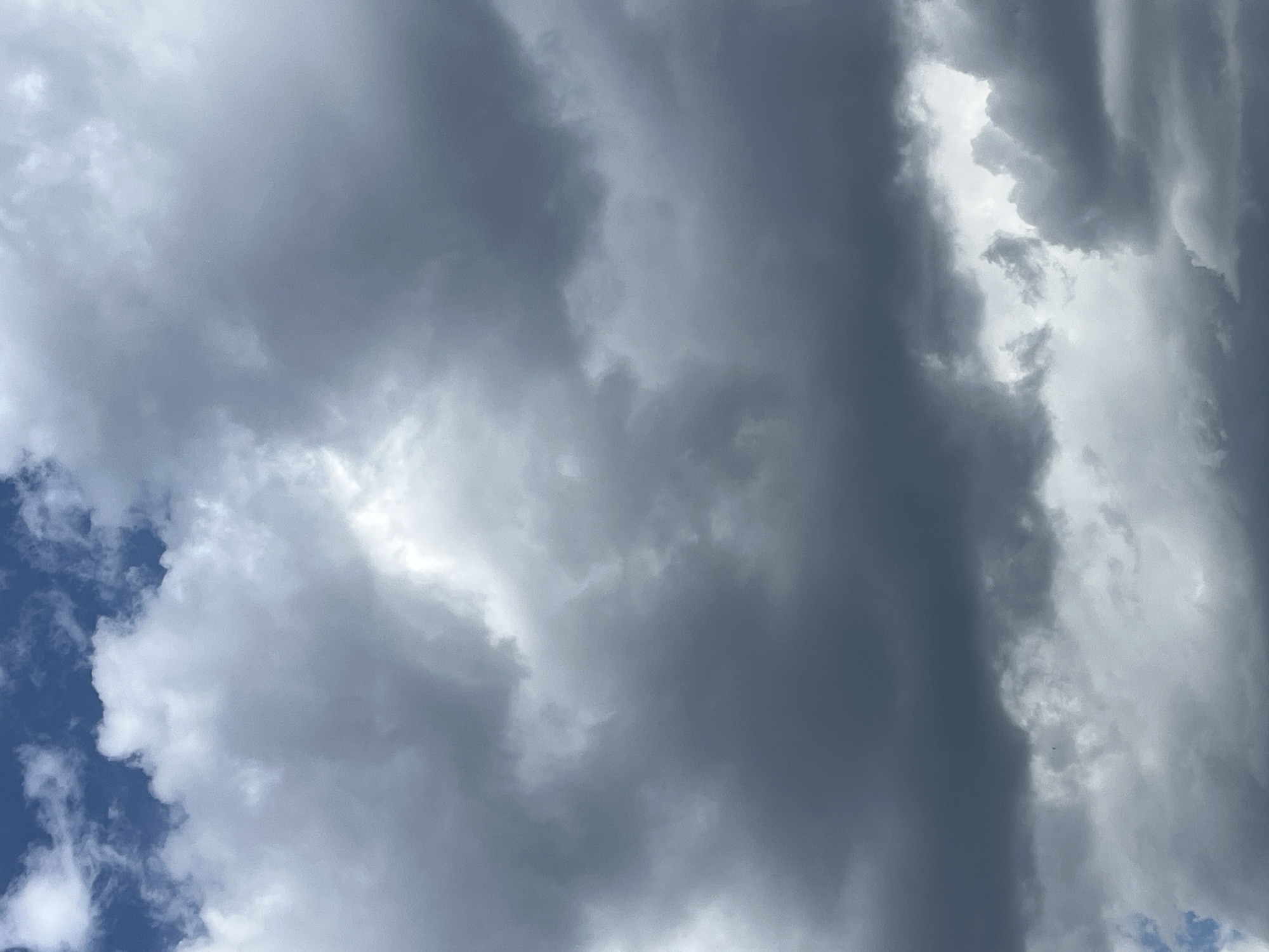 This media file is a photograph of a cloudy sky. The clouds are varied in color and texture, ranging from dark gray to bright white, suggesting a mix of heavy and light cloud cover. There are also patches of blue sky visible, particularly in the lower left corner, indicating that it is daytime. The lighting suggests that the sun is either obscured by clouds or low in the sky, potentially indicating early morning or late afternoon. The overall impression is one of dramatic and dynamic atmospheric conditions. There are no visible people, objects, text, or specific location cues to provide further context.