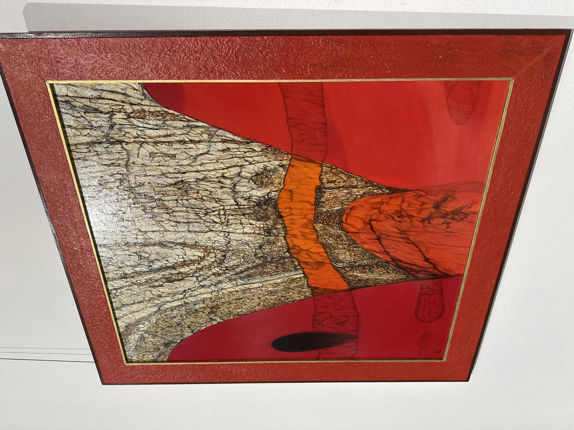 This media file captures an abstract framed artwork displayed on a white wall in an interior setting, likely in Minsk, Belarus. The artwork itself is a mixed-media piece, dominated by contrasting colors and textures.
The left side of the artwork features a prominent, highly textured area resembling cracked earth, aged parchment, or a dried-out landscape, rendered in shades of grey, beige, and black with intricate, fine dark lines forming a web-like pattern. This textured section sharply abuts large, smooth expanses of vibrant red. Integrating these sections are dynamic, elongated forms in an organic orange hue, characterized by a fibrous or streaky texture, reminiscent of tree bark or flowing fabric. One such orange form extends vertically near the center, while others are seen towards the right.
A notable detail in the lower red section is a distinct, elongated black oval, which could be interpreted as a shadow or an abstract element within the composition. The artwork is enclosed within a striking red-orange frame, which also exhibits a textured finish, possibly mimicking leather or a similar material. An inner thin gold border separates the artwork from its frame. The image is captured from an upward, angled perspective, making the frame appear trapezoidal. In the bottom right corner of the artwork, within a red area, a small, faint, and stylized signature or symbol is barely discernible.