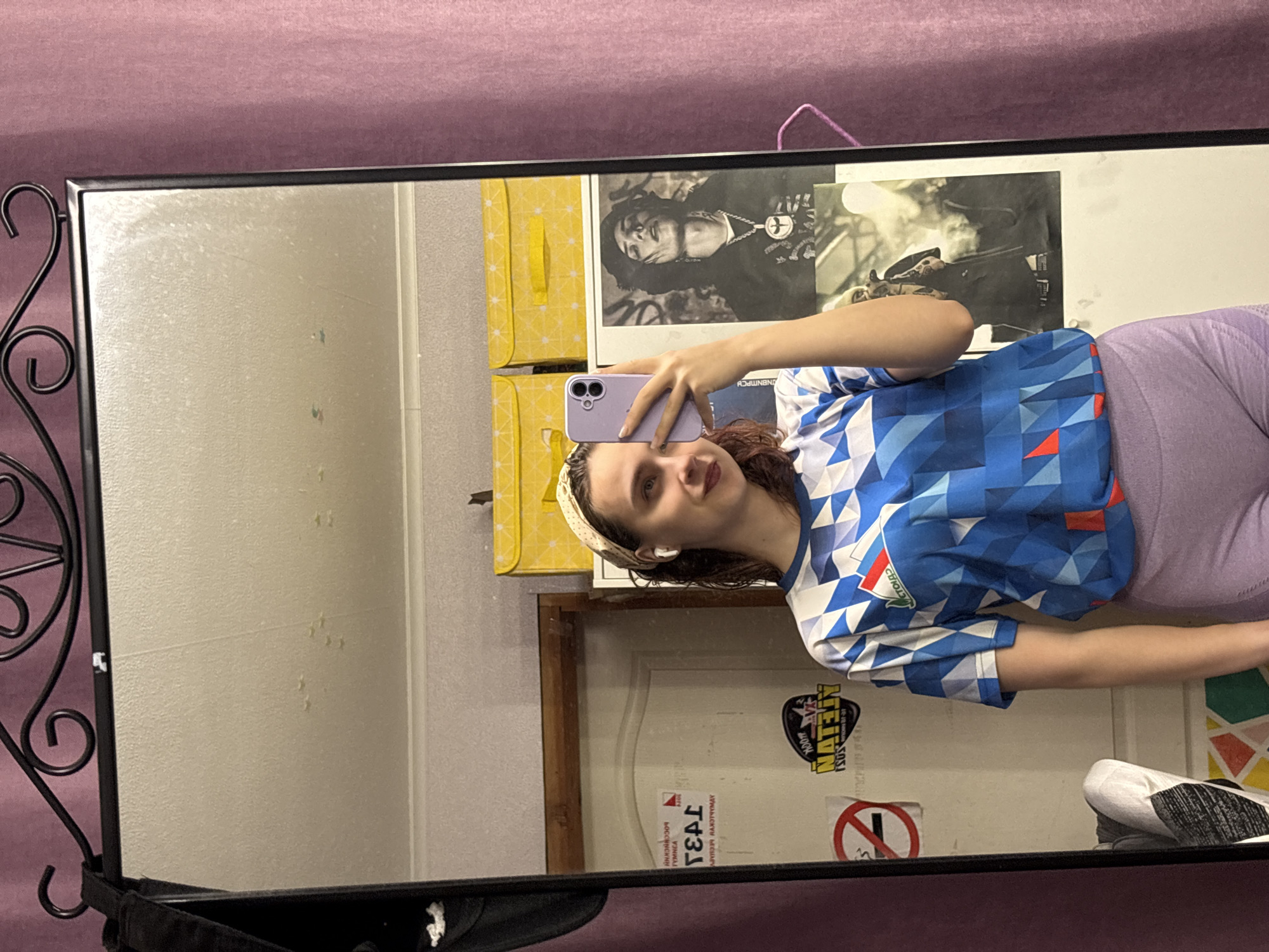 A woman is taking a selfie in a mirror. The mirror has a black wrought iron frame. The woman is wearing a blue and white geometric pattern shirt and light purple leggings. She is holding a light purple smartphone. Behind her are posters, a yellow storage box, and a door with stickers. The location appears to be indoors. The city of Прикамский, Russia, cannot be determined from the image.