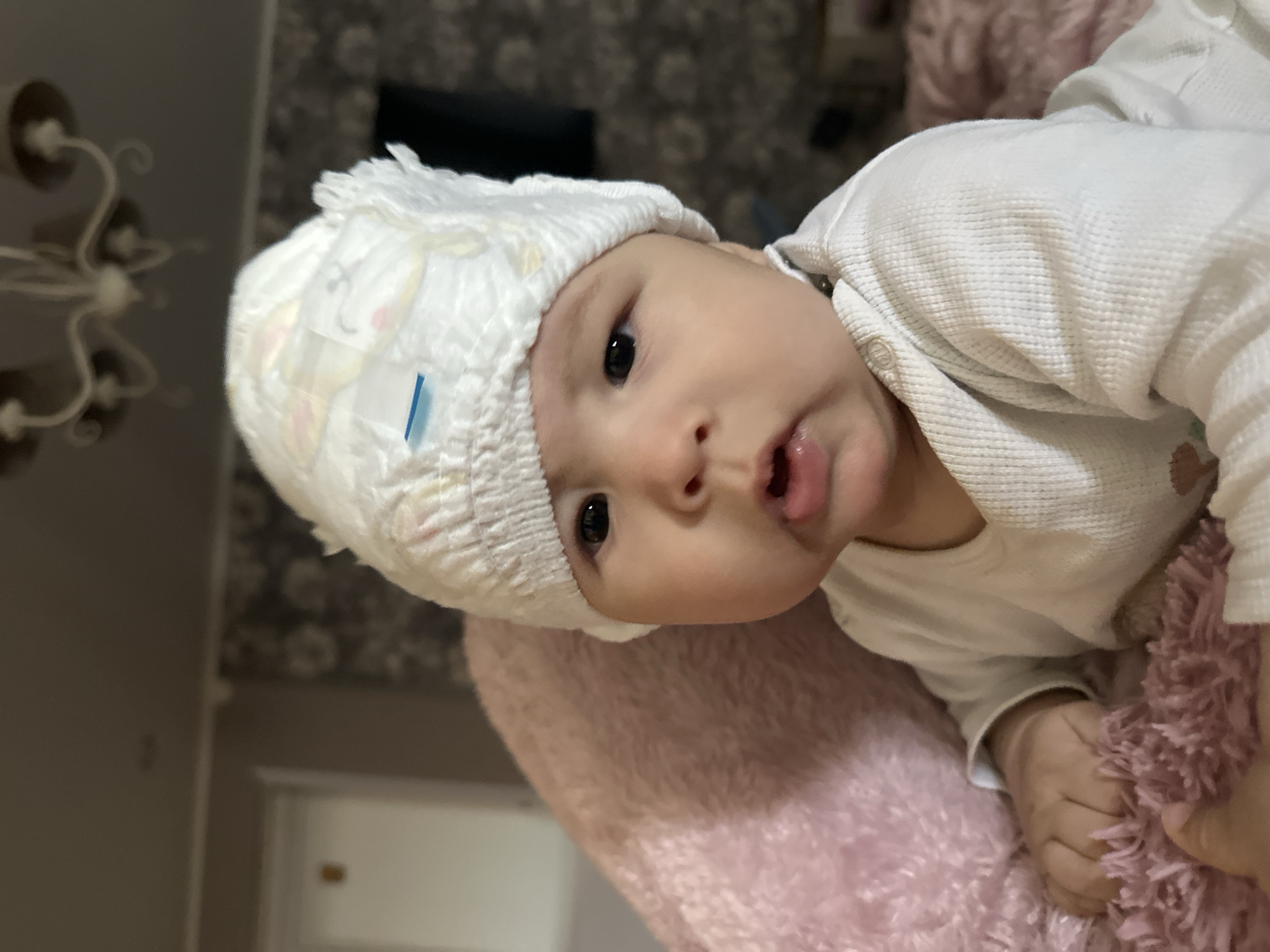 A baby is lying on a pink, furry surface. The baby is wearing a white, long-sleeved shirt and a diaper as a hat. The background includes a patterned wall and a light fixture. The location appears to be indoors, possibly a bedroom or living area.