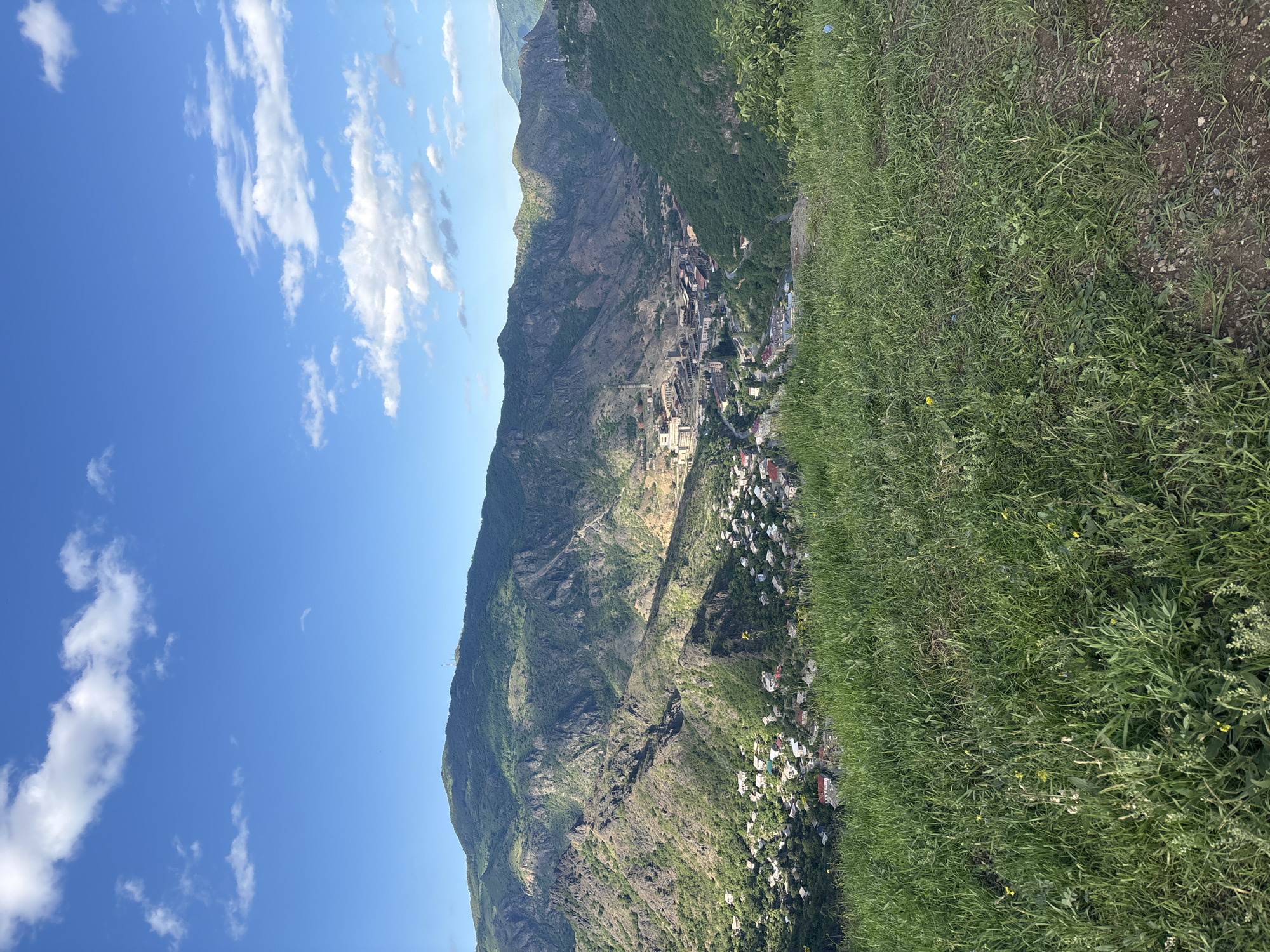 A landscape view depicts a mountain range with a town nestled on its slopes under a blue sky with scattered white clouds. The foreground features green vegetation and some bare earth, suggesting an elevated observation point. The mountain range is covered in green vegetation and has patches of exposed rock. The town is comprised of buildings of varying sizes and colors, primarily clustered together. The sky is blue with puffy white clouds filling part of the frame. The scene appears to be captured during daylight, possibly late morning or early afternoon. The location is Akori, Armenia.