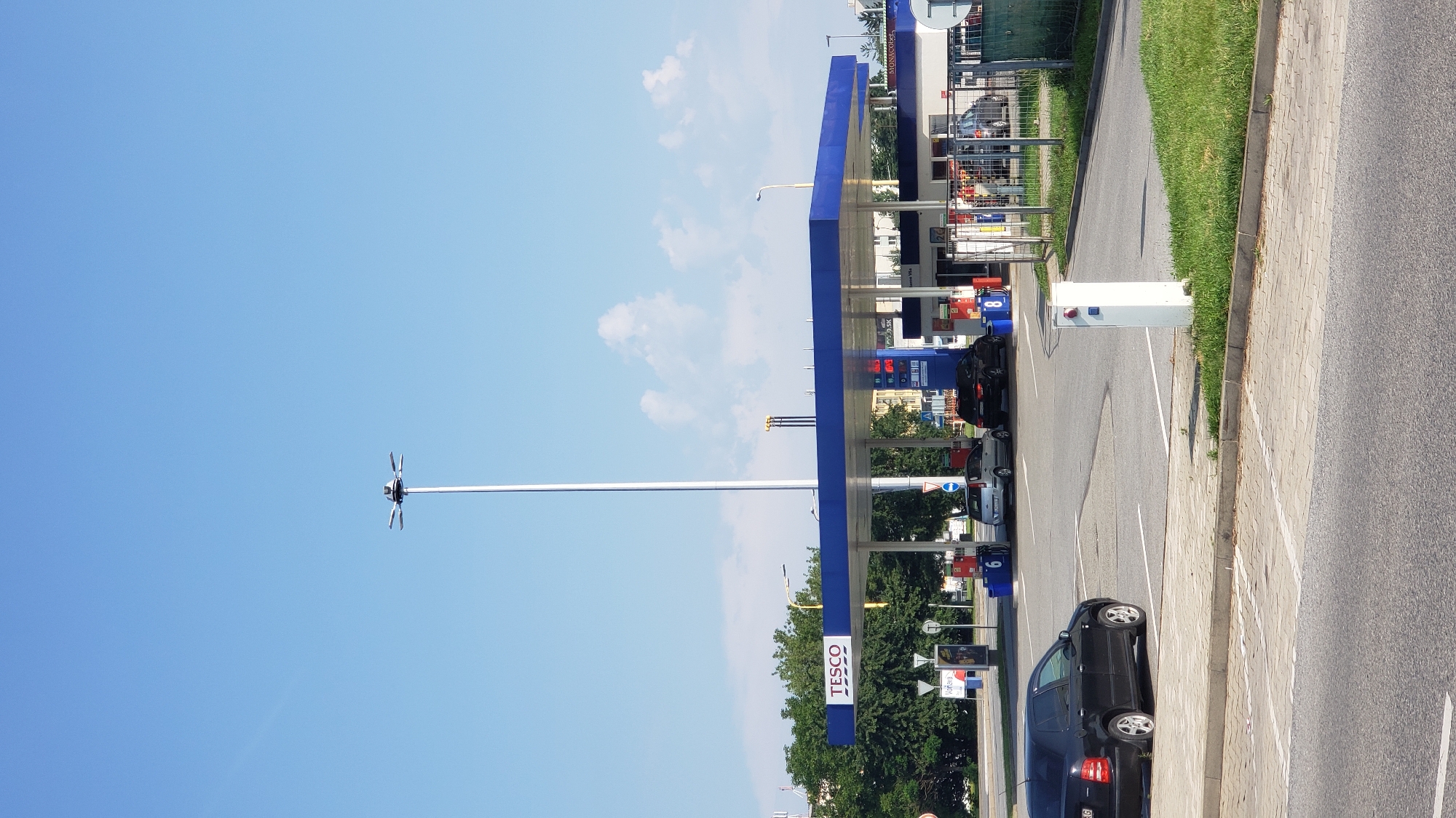 The image shows a Tesco petrol station located in Prešov, Slovakia. A tall, multi-armed lighting mast stands in the background, with its base obscured by the petrol station canopy. The canopy, painted in blue and white, covers multiple fuel pumps. Several cars are visible, including a black Skoda Fabia parked in the foreground, a silver car at one of the pumps, and two other cars further back. A white payment kiosk with blue and red buttons is positioned near the foreground parking area. The environment is characterized by a clear blue sky with scattered clouds, green trees lining the perimeter of the station, and paved surfaces for the forecourt and parking.