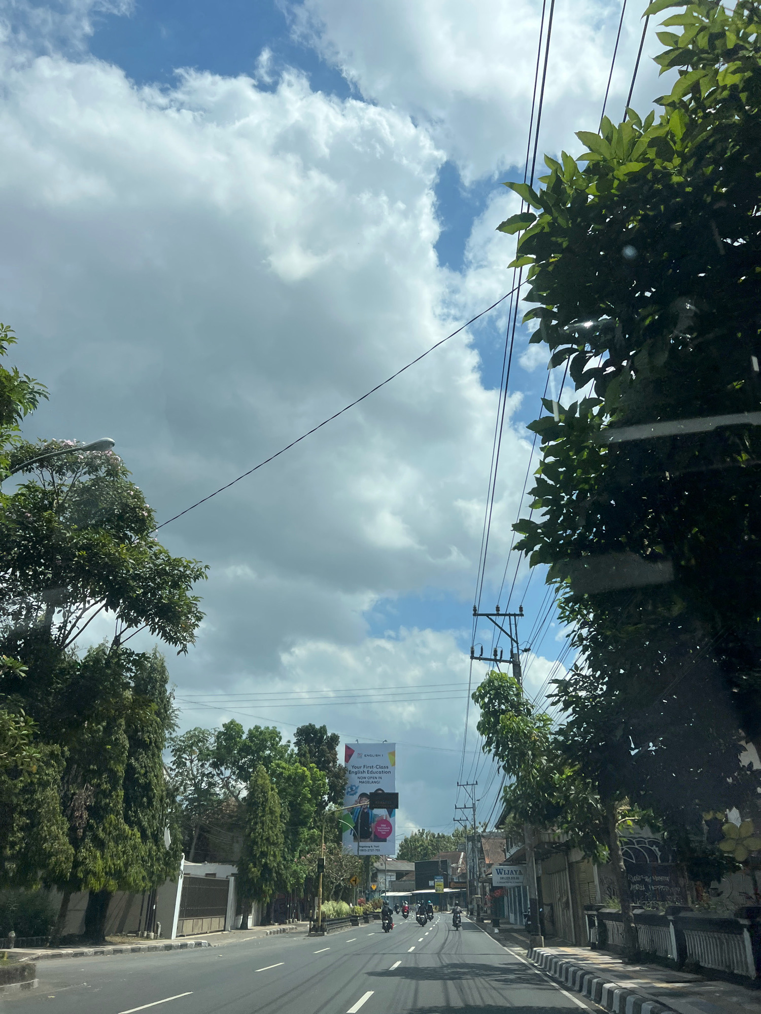 This is a vertical, eye-level, full shot of a street in the city of Magelang, Indonesia. The sky is mostly cloudy with patches of blue. There are power lines running across the frame. Trees line both sides of the street, with the foliage on the right being denser and closer to the camera.
On the left side of the street, there are several tall, slender evergreen trees and a few wider, fuller trees. Behind them, a white building with a light-colored fence is visible. Further down the road, a large billboard with the English text "Your First-Class English Education" and the location "MAGELANG" is displayed.
On the right side of the street, there are buildings with storefronts. One sign reads "Wijaya" and displays a phone number. The buildings have a more traditional architectural style. There are also smaller trees and a black and white striped wall along the sidewalk.
The road is asphalt and has white lane markings. Several motorcycles are being ridden down the street, moving away from the camera. The overall impression is a typical street scene in a developing city.
