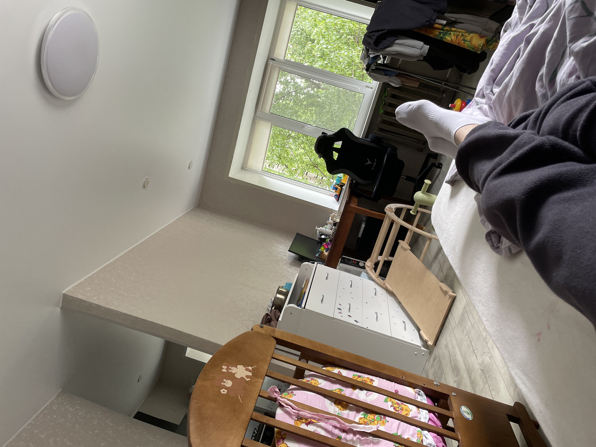 A skewed, high-angle indoor shot shows part of a room in Perm, Russia. Dominating the right side of the frame is a person on a bed, partially visible from the legs down, wearing white socks and dark sweatpants. The bed is covered with white bedding.
In the background, a window is visible, showing green foliage outside. Near the window is a black gaming chair, and next to it, a messy shelf with clothes.
In the lower left of the frame, a wooden crib with pink patterned bedding is situated near a white dresser with numbered drawer pulls.
On the ceiling, a round, white light fixture is visible. The room's wall has a light-colored patterned wallpaper. The viewpoint suggests the photo was taken from an elevated position, possibly a bed or another high surface.