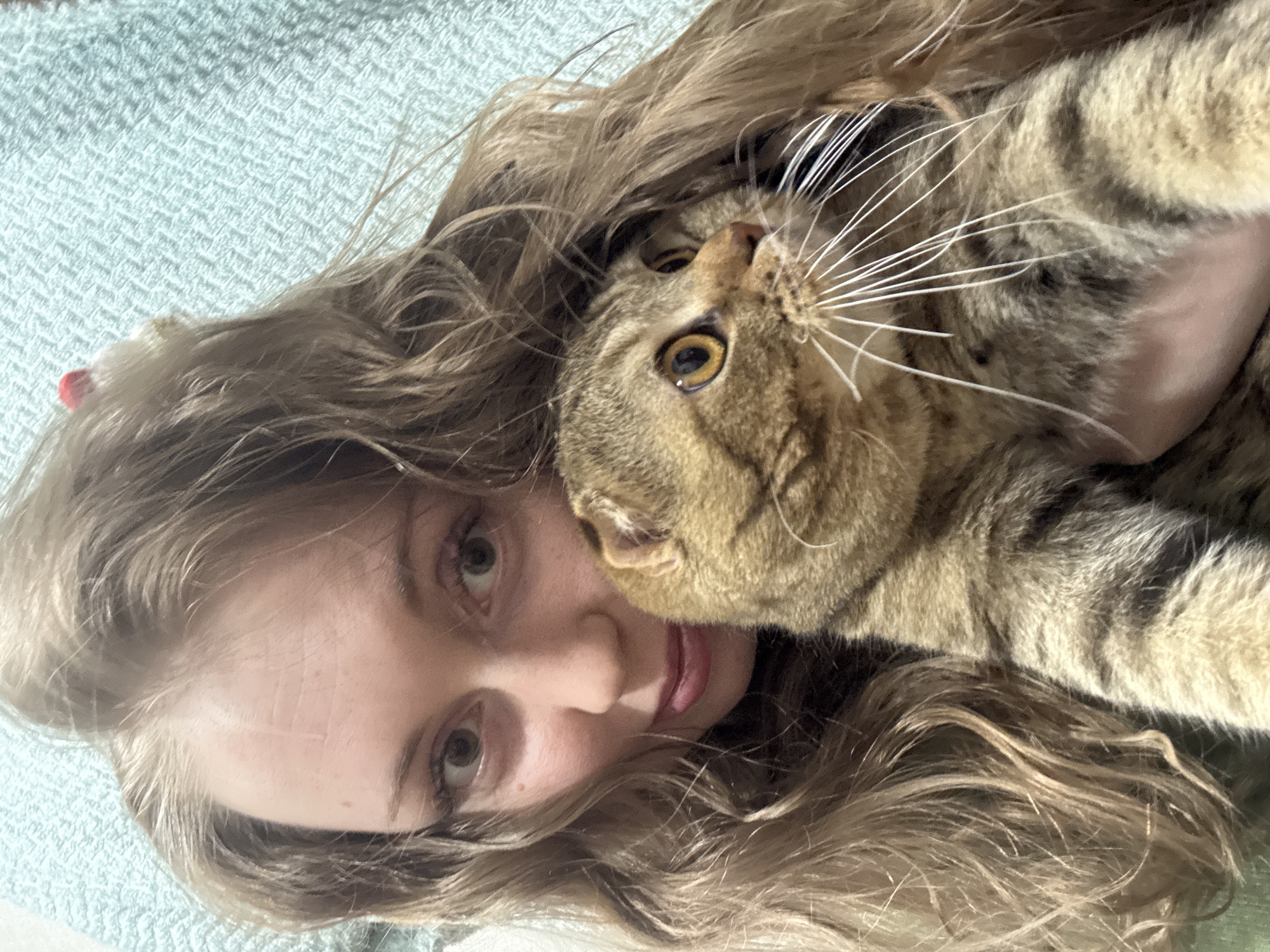 A woman with long, wavy brown hair is lying on a light blue textured surface, possibly a bed or couch. She is holding a brown tabby cat. The cat is positioned so its head is near the woman's face, and its long white whiskers are prominent. The cat is looking upwards. The woman is looking at the camera.