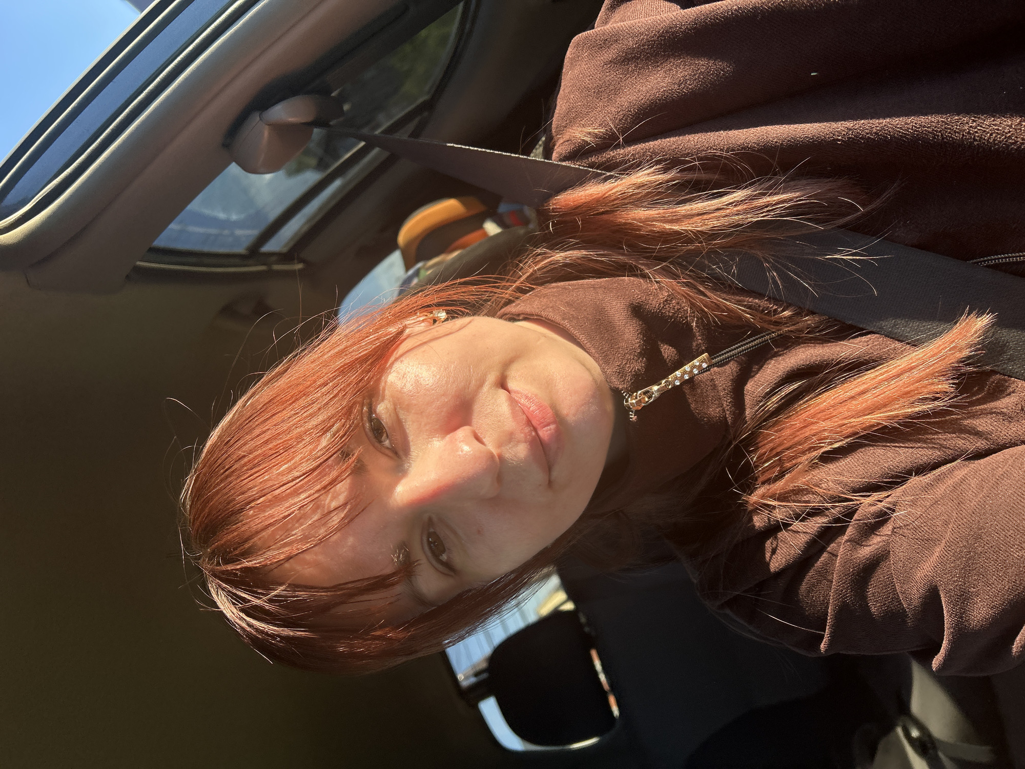A woman with reddish-brown hair and wearing a brown jacket is sitting in a car. She is wearing a seatbelt. The car has a sunroof visible in the upper left. The photo was taken inside the car, looking up and slightly to the right at the woman. The location is likely not Фоки, Russia based on the information in the photo.