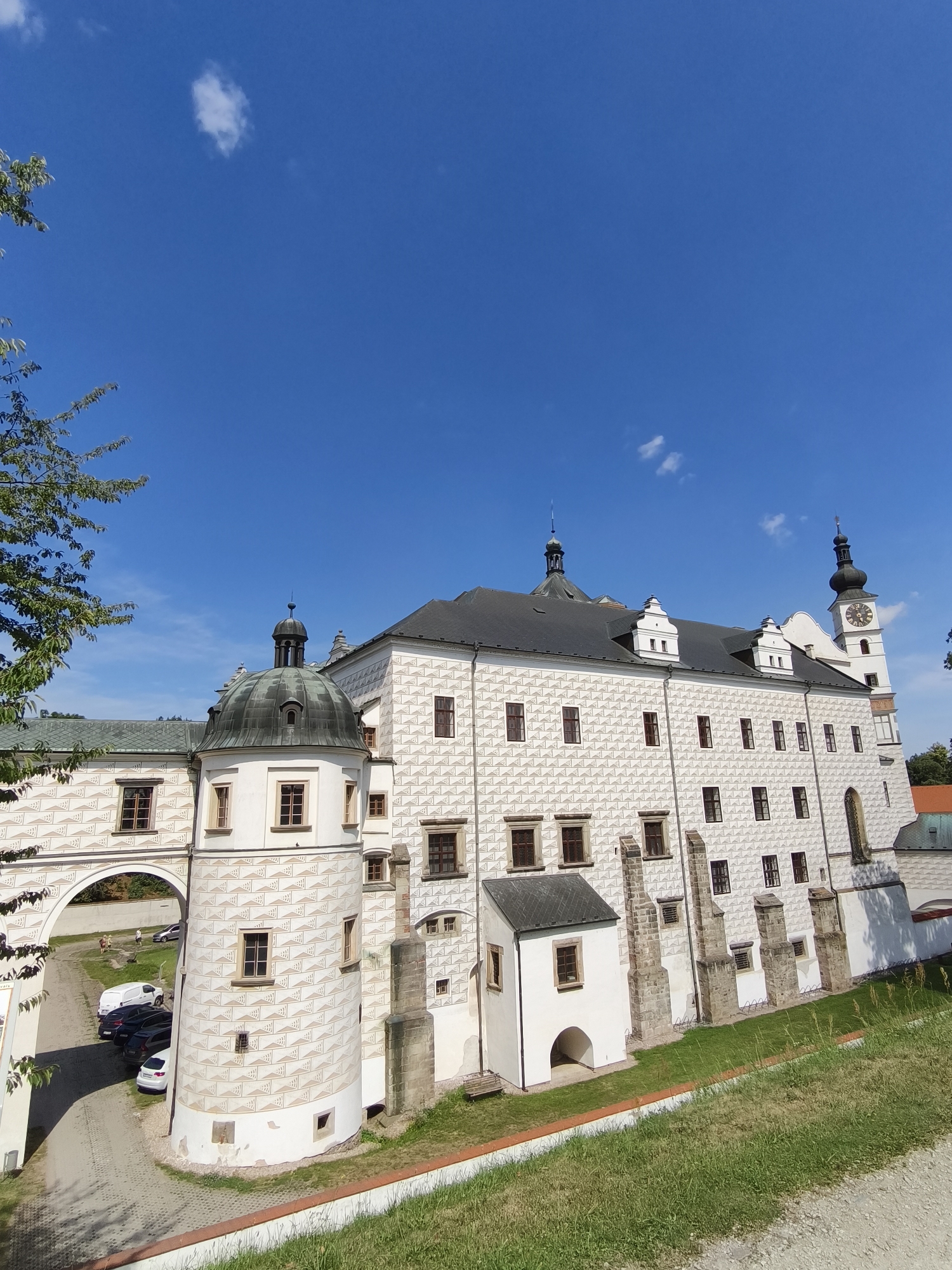 The image shows a large, white, ornate building with a dark roof, possibly a castle or palace, under a clear blue sky with a few scattered clouds. The building features numerous windows, decorative patterns on its facade, and several architectural elements including a round tower with a domed roof and a taller tower with a clock face. In the foreground, there is a grassy slope and a paved area with several parked cars. To the left of the building, an arched walkway connects to another part of the structure. The style of architecture suggests a historical European location. This building is the Pardubice Castle, located in the city of Pardubice, Czechia. The castle is a prominent landmark and a popular tourist destination, known for its Renaissance architecture and expansive courtyards.