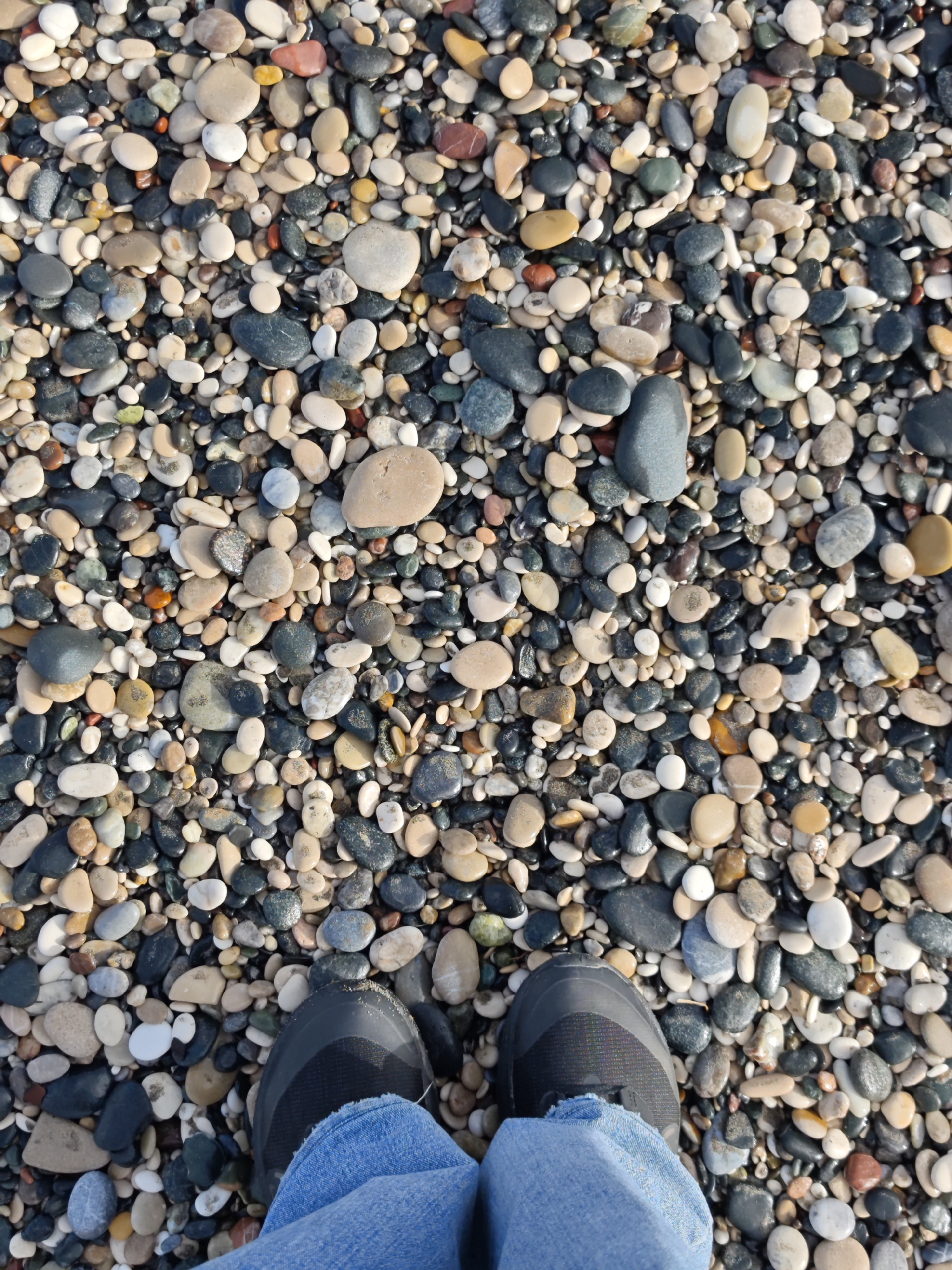 The image depicts a ground-level view of a pebble beach in Demre, Turkey. The scene is dominated by an assortment of small, smooth, rounded pebbles of varying colors, including white, beige, brown, gray, and black. Interspersed are also a few colorful orange and green pebbles. In the bottom portion of the image, there are two feet, wearing dark-colored sneakers and light blue denim jeans. The shoes are partially embedded in the pebbles. No other individuals or objects are discernible.