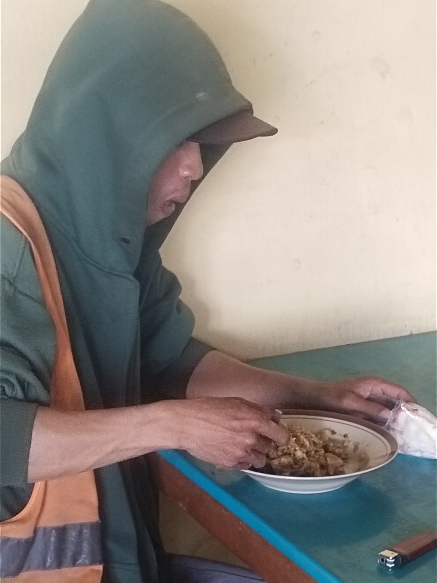 A person wearing a green hooded jacket and an orange vest is sitting at a table in Kemlakagede, Indonesia, eating from a bowl. The person is using their hands to eat the food, which appears to be rice and some other ingredients. A lighter and a plastic bag of food are also on the table. The wall in the background is a light color. The overall scene suggests a casual meal being eaten by an individual.
