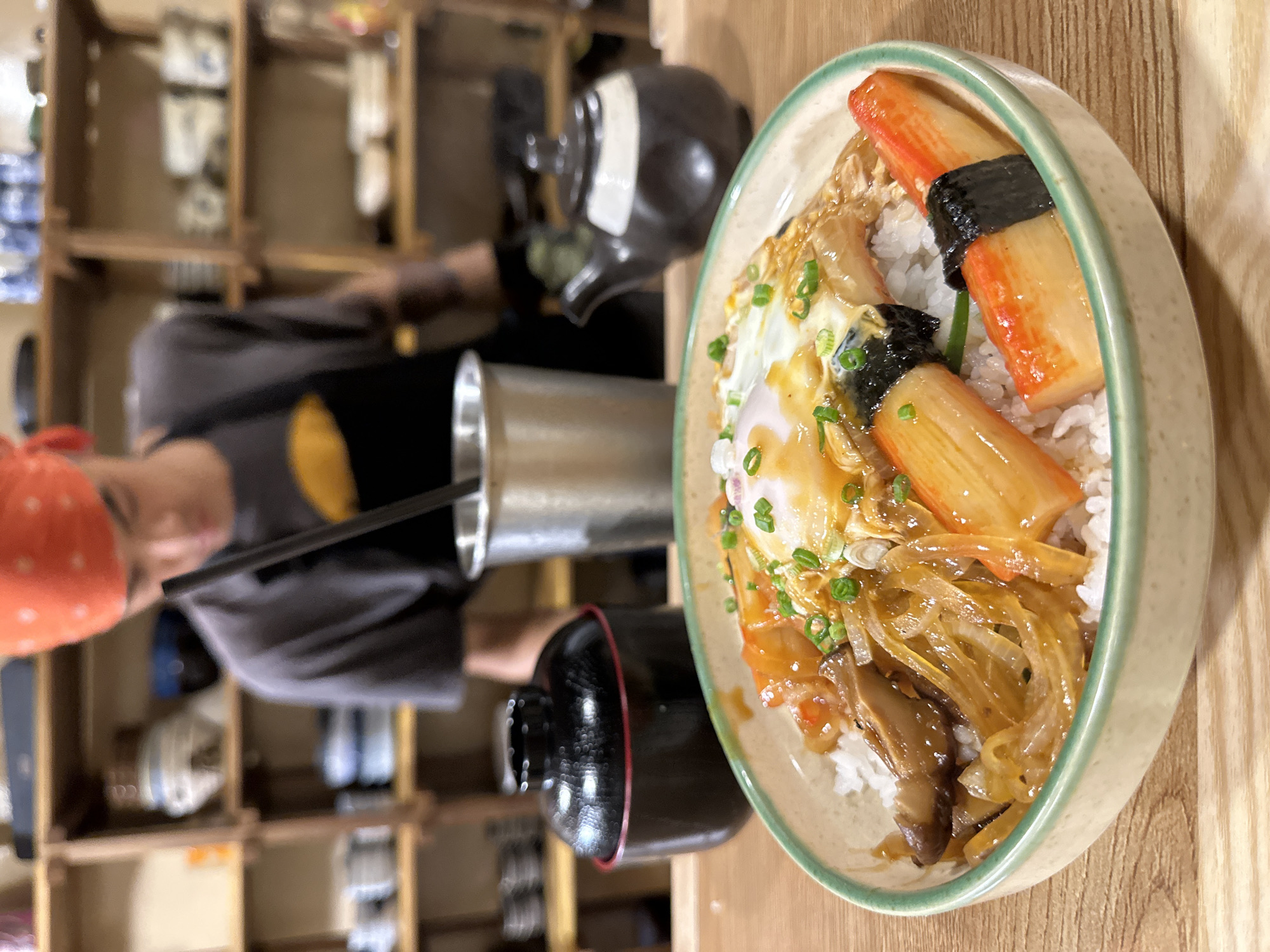 In the foreground, a bowl of rice is filled with various toppings, including imitation crab sticks, mushrooms, onions, and a fried egg. The bowl has a light green rim and rests on a wooden table. To the left of the bowl is a black lidded container with a red rim. Behind the bowl, a person wearing an orange bandana and a grey shirt is visible, looking down and stirring something in a metal cup. Shelves filled with ceramic dishes are arranged in the background. To the right of the metal cup, there is a small, dark brown and white teapot. The scene appears to be in a restaurant setting, possibly in Da Lat, Vietnam, given the style of the dishes and the presence of a person in what seems to be a kitchen or food preparation area.