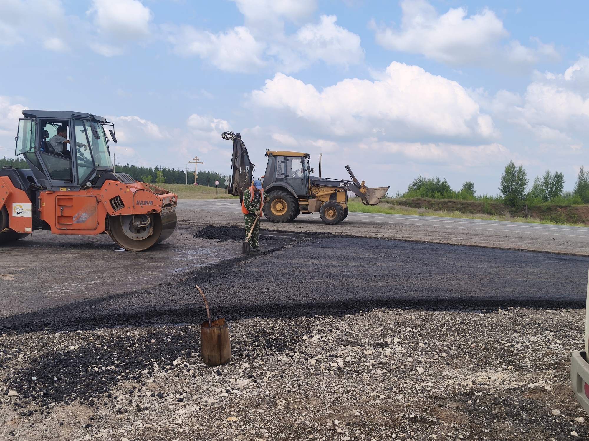 The image shows a road construction scene on a sunny day. In the foreground, a roller is compacting freshly laid asphalt. A worker in a camouflage uniform and a high-visibility vest is using a shovel to spread the asphalt. Behind them, a yellow backhoe is parked, with its bucket raised. The background features a road, trees, and a cloudy blue sky. In the distance, there appears to be a wooden cross on a small hill. This scene is located in Borskoe, Russia.