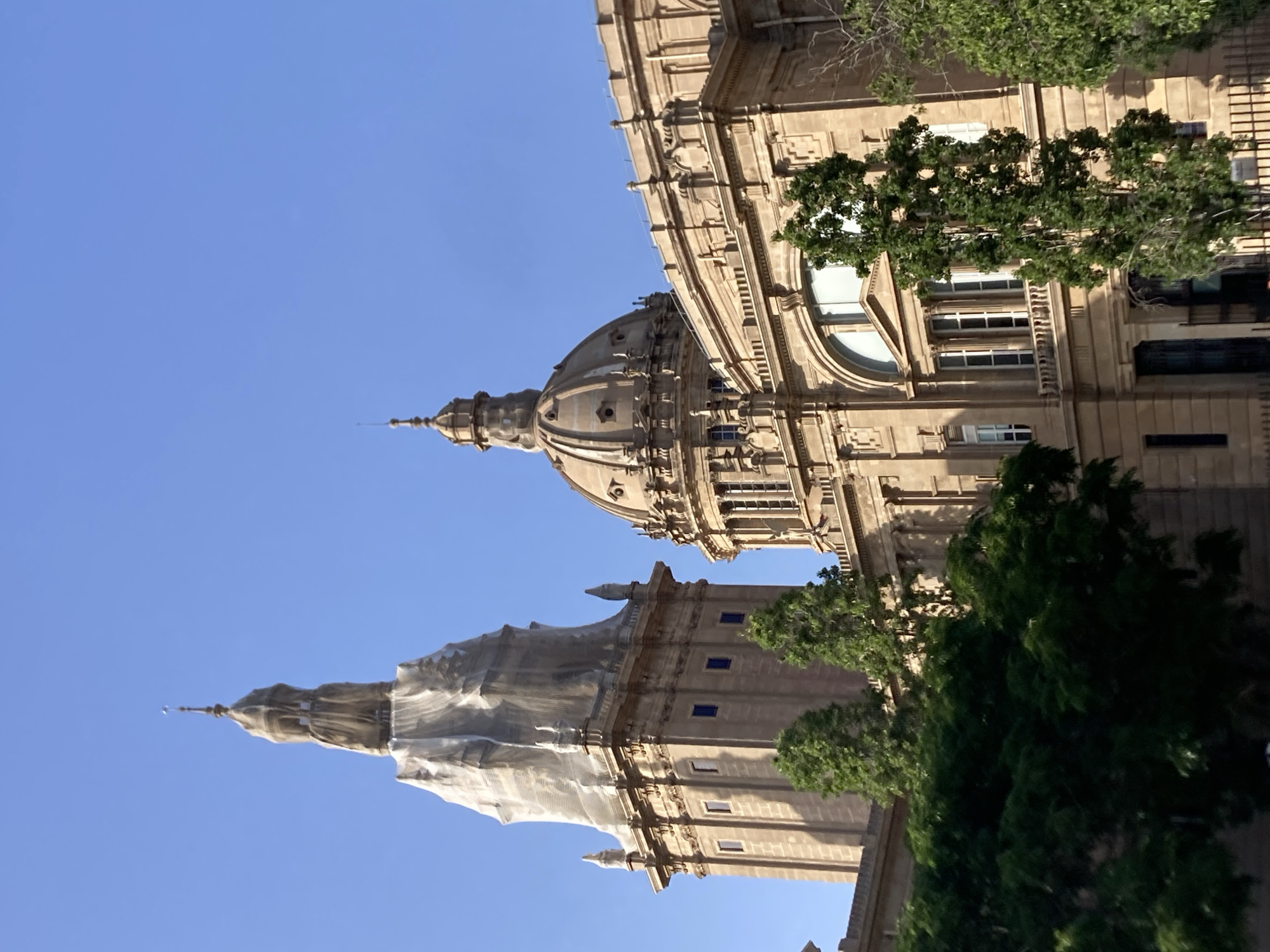 The image captures a low-angle view of the Palau Nacional, a majestic palace located on Montjuïc hill in Barcelona, Spain. The palace, constructed of stone, features classical architectural elements, including a prominent dome and a tall tower. The tower on the left is partially covered in white scaffolding, suggesting ongoing restoration or maintenance work. Green trees are visible in the foreground, partially obscuring the lower levels of the building. The sky is clear and blue, indicating a daytime scene. The architecture suggests a grand, historical building, likely a museum or cultural institution.