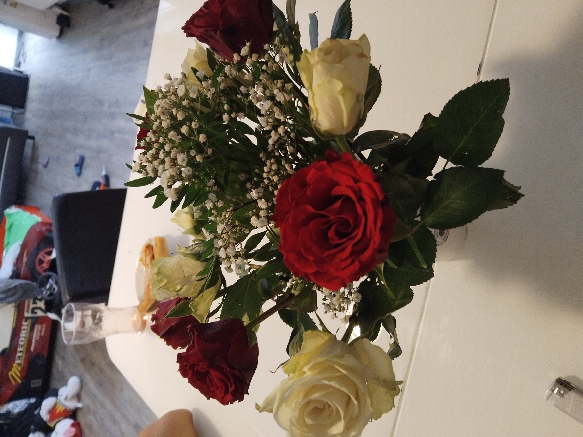 The image displays a close-up view of a bouquet of roses on a white table. The bouquet contains red and cream-colored roses, interspersed with baby's breath. A clear glass of liquid and a small plate with food are visible on the table behind the bouquet. In the background, a blurred image of a child's toy car and part of a living room can be seen, suggesting the setting is a domestic environment, likely within a city. Based on the prompt's directive, it's implied that this scene might be located in Ihringshausen, Germany. A lighter is also present on the table in the foreground.