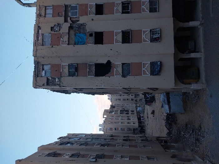 The media file displays a scene of widespread destruction in an urban environment, consistent with the provided context of Khan Yunis, Palestinian Territory. The image is oriented vertically but depicts buildings standing upright, suggesting a landscape of multi-story structures.
The primary subjects are several heavily damaged buildings, likely residential or commercial. The building in the foreground, and others visible in the background, exhibit extensive structural damage. Many windows are completely blown out, leaving dark, empty rectangular openings. Some window frames remain, but the glass is shattered or missing. Visible scorch marks blacken parts of the facades around these openings, indicative of fires or explosions. Sections of the buildings' outer walls are collapsed or crumbled, exposing interiors and structural elements like rebar. On one building, a distinct blue tarp is visible, covering a significant opening or damaged section of a wall, possibly as a makeshift repair or protection. The buildings originally appear to have been light-colored with decorative surrounds around their windows.
The ground level between the buildings is covered in a substantial amount of debris, rubble, and dirt. This area looks like a barren wasteland, further emphasizing the devastation. Dark shapes possibly indicating damaged or abandoned vehicles are visible in the shadowy areas near the base of one building.
The scene takes place during daylight hours, under a clear sky, highlighting the extensive damage. There are no people visible, and no identifiable text on any of the structures or debris. The overall atmosphere conveys desolation and the profound aftermath of conflict or disaster.