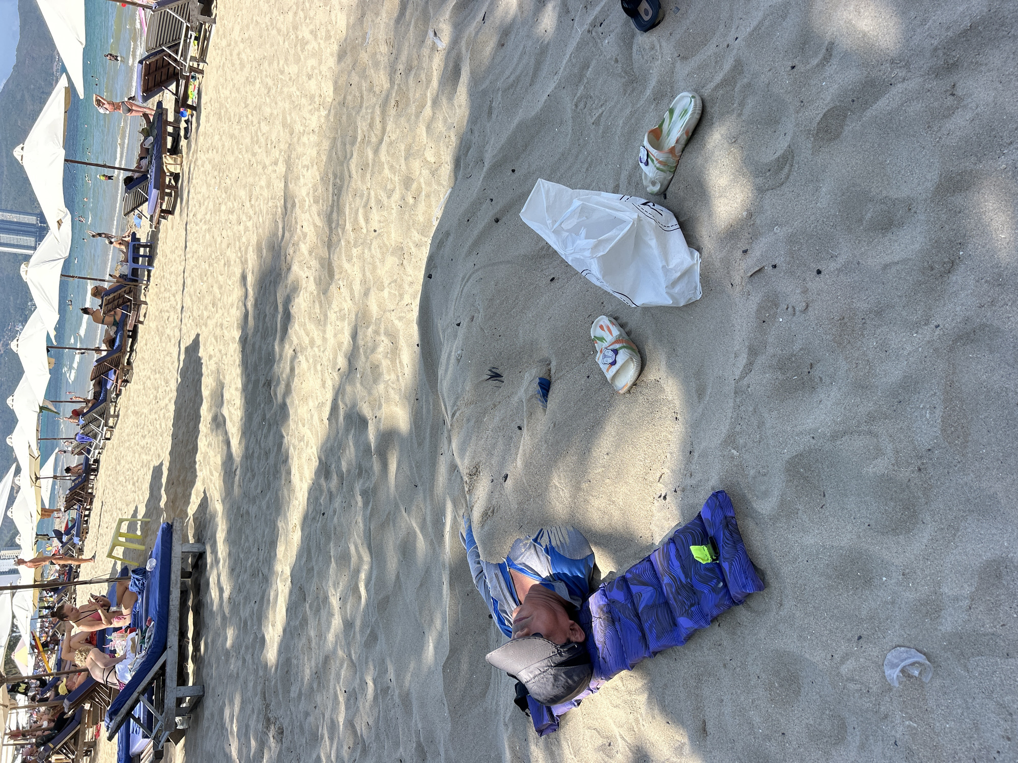 The image depicts a scene on a sandy beach, likely in Bac Nha Trang Ward, Vietnam, given the presence of high-rise buildings in the background and the beachside setting. A man is lying on a blue and black patterned sleeping pad in the sand. He is wearing a grey cap, a light blue shirt, and darker shorts. Near him are a pair of patterned sandals, a plastic shopping bag, and what appears to be a small piece of plastic litter. In the background, there are beach umbrellas, lounge chairs, and people, suggesting a resort or public beach. The overall scene is outdoors, during daylight, with sunlight casting shadows across the sand. The composition suggests an area where leisure and tourism activities take place.