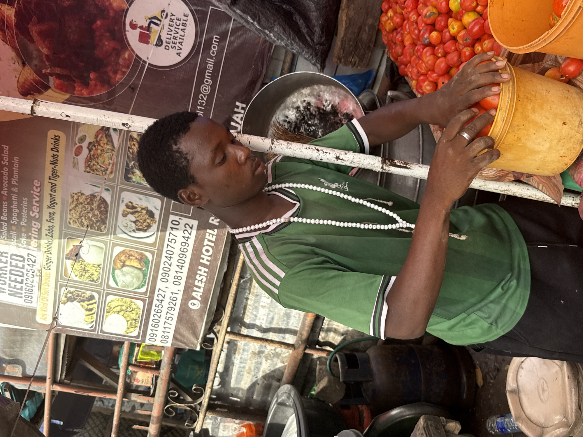 The image, set in Lekki, Nigeria, captures a young man working at what appears to be an outdoor food stall or market stand. He is focused on handling fresh red tomatoes, transferring them between orange plastic buckets. He wears a green short-sleeved shirt with white and black stripes on the sleeves and collar, black shorts, a white beaded necklace, and a silver ring on his left hand. His expression is neutral and concentrated on the task.
The stall is equipped with large cooking pots and possibly gas cylinders, suggesting food preparation or sale. Behind and to the side of the man are several signboards displaying various food items and services. One large sign advertises a menu including "Salad Beans Avocado Salad," "Macali Spaghetti & Plantain," "Cake Pastries," and "Any Kind Of Soft Drinks: Ginger Drinks, Zobo, Fura, Yogurt and Tiger-Nuts Drinks." This sign also states "DELIVERY SERVICE AVAILABLE" and lists contact information including multiple phone numbers: "09160285427," "09160265457," "09024075710," "08117579261," "08140969422," and an email address "132@gmail.com." Another section of the sign indicates "WORKER NEEDED" and the partial name "ALESH HOTEL R," likely referring to a nearby establishment or an associated catering service. The word "JAH" is also visible on another sign fragment.
Piles of bright red tomatoes are prominent, some already in buckets, others spilling from larger containers, indicating a fresh produce supply. The overall scene suggests a vibrant, busy commercial environment focused on food sales and catering services in a local setting, likely during daytime.