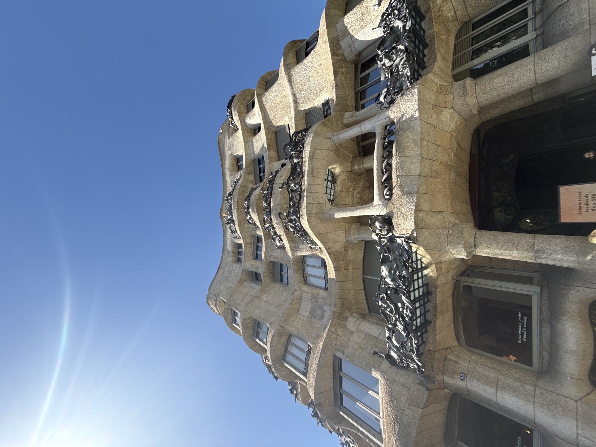 The image shows a low-angle shot of the exterior of Casa Batlló, a famous building designed by Antoni Gaudí, located on Passeig de Gràcia in Barcelona, Spain. The building's facade is characterized by its undulating, organic shapes, reminiscent of bone and wave forms, constructed from stone and featuring irregularly shaped windows and balconies with wrought-iron railings. The sky is a clear, bright blue with a visible lens flare from the sun. The sunlight creates a strong contrast between the illuminated parts of the building and the shadowed areas. Several windows are visible, some with shutters, and some reflecting the sky. Text is visible on some windows, including "Gastronomy under Gaudí's ceiling." A sign with Spanish text is also partially visible at the bottom. There are no people clearly identifiable in the image.