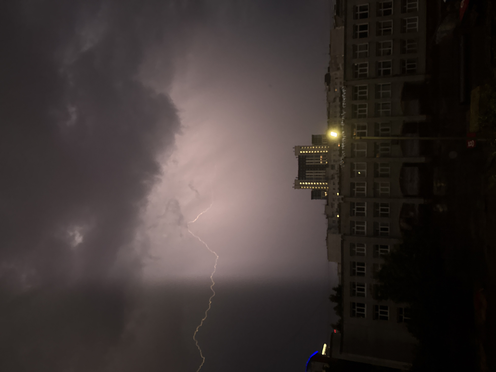 An outdoor, low-angle, vertical shot shows a dramatic lightning strike illuminating a dark, stormy sky over a city. The lightning bolts, jagged and bright yellow, stretch across the upper left quadrant of the frame, cutting through heavy, dark grey storm clouds. The contrast between the dark clouds on the left and the lighter, pinkish-purple sky illuminated by the lightning on the right is stark.
In the mid-ground, a long, multi-story building with many rectangular windows is visible. It appears to be an institutional building, possibly a school or dormitory. A street light with a bright yellow bulb shines in front of this building, casting a warm glow. Behind this, a taller, modern building with many illuminated windows stands out against the stormy sky, its lights creating a grid pattern. The upper portions of these buildings are obscured by the dark clouds and atmospheric haze.
The foreground is predominantly dark, with the silhouettes of trees and possibly other structures hinting at ground level. The overall scene conveys a sense of powerful natural phenomena occurring in an urban setting.