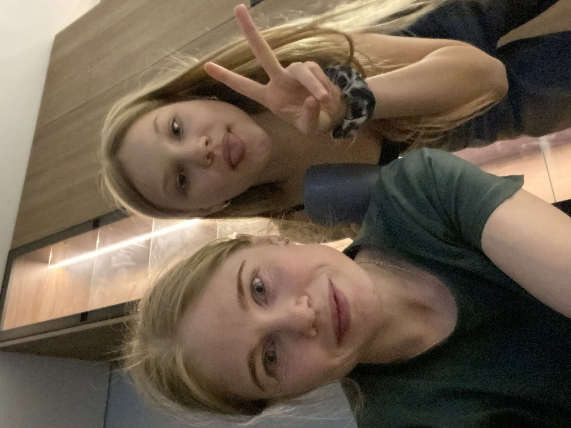 Two young women are posing for a selfie. The woman in the foreground has blond hair and is wearing a dark green t-shirt and a necklace. The woman in the background has light blond hair, is sticking her tongue out, and making a peace sign with her right hand. She has a scrunchie on her wrist. They appear to be indoors. Behind them is a wooden cabinet with built-in lighting. It is not possible to determine the location in the city of Chaikovsky, Russia, based on the provided image.