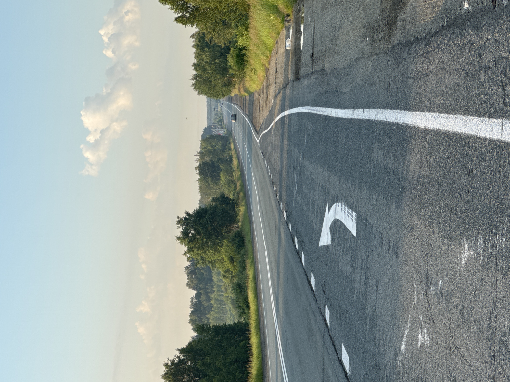 A vertical, eye-level shot captures an asphalt road in a rural setting under a clear sky. The road is divided by white lines, with a large white arrow pointing left painted on the right lane. Trees line both sides of the road, with a dense forest in the background. A few cars are visible in the distance, and the sky is a pale blue with scattered white clouds. The overall impression is one of a serene, rural landscape. The location is identified as Unknown, Russia.