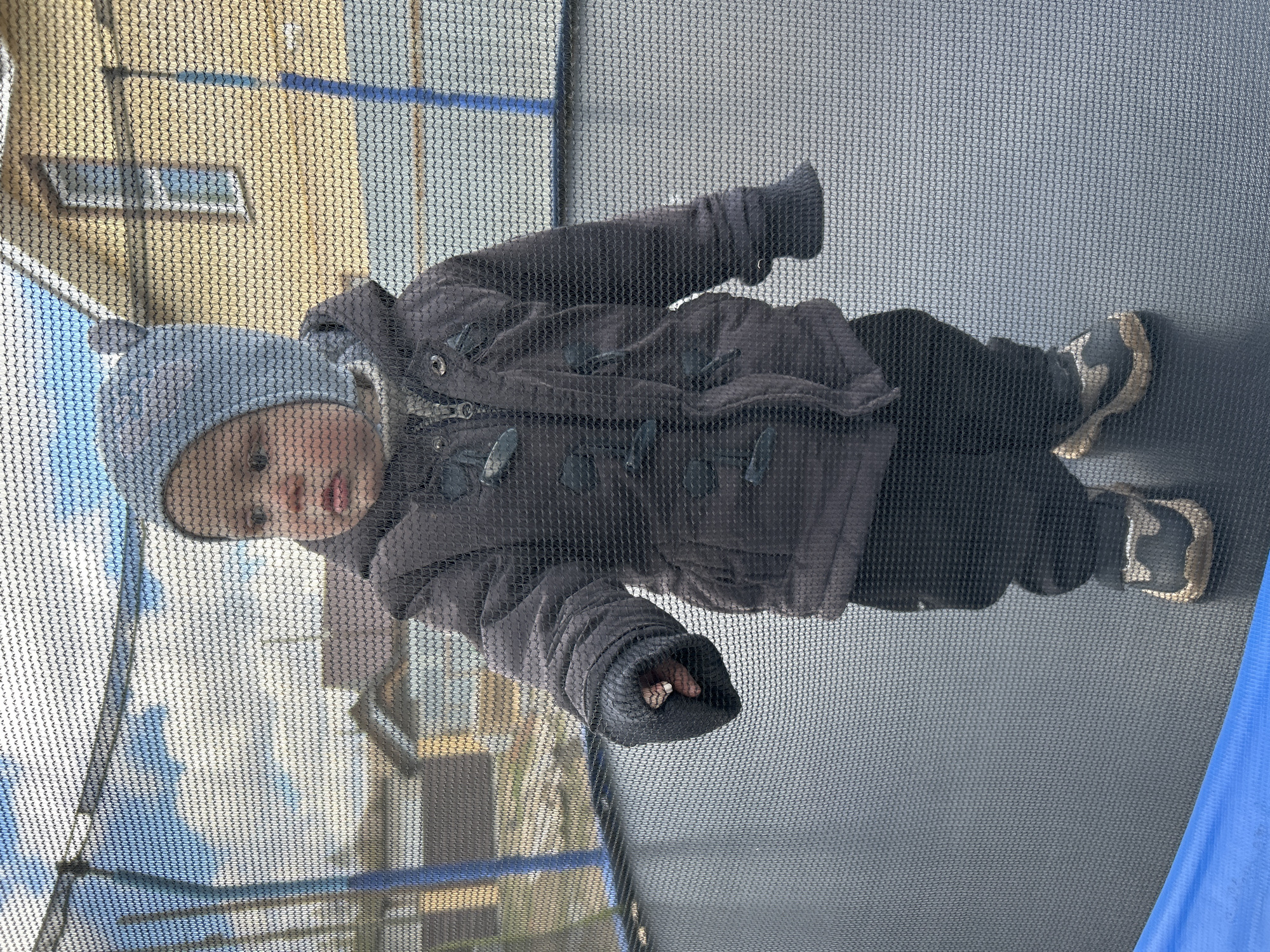 A toddler wearing a gray hat, a dark gray jacket, and dark pants is standing on a gray trampoline enclosed by a black net. The trampoline has blue padding on one side. The toddler is facing the camera. A building with a window is visible through the netting in the background. The image was taken in Chaikovsky, Russia.