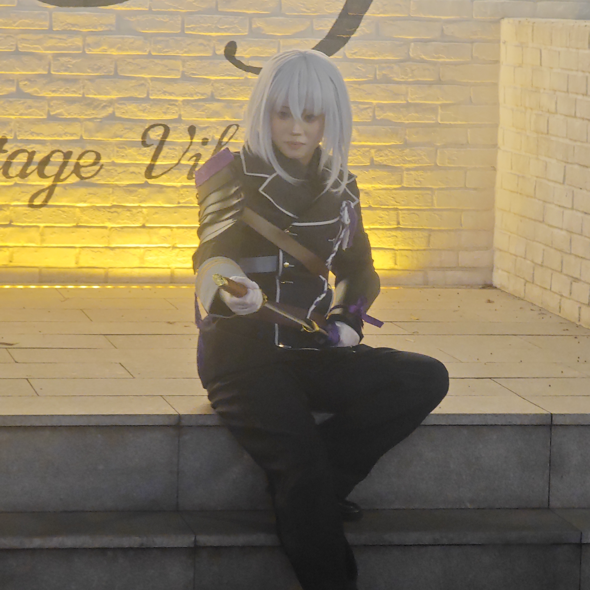 A person in cosplay sits on a set of stone steps. The person has short white hair and is wearing a black uniform-style jacket with gold and purple accents, as well as black pants. They are holding a sheathed sword in their right hand. The background consists of a wall constructed of light-colored bricks. A word is partially visible on the wall, and illuminated by yellow light. The location is most likely a staged background in an urban area, but the specific location in Liangjiang, China is not discernable.